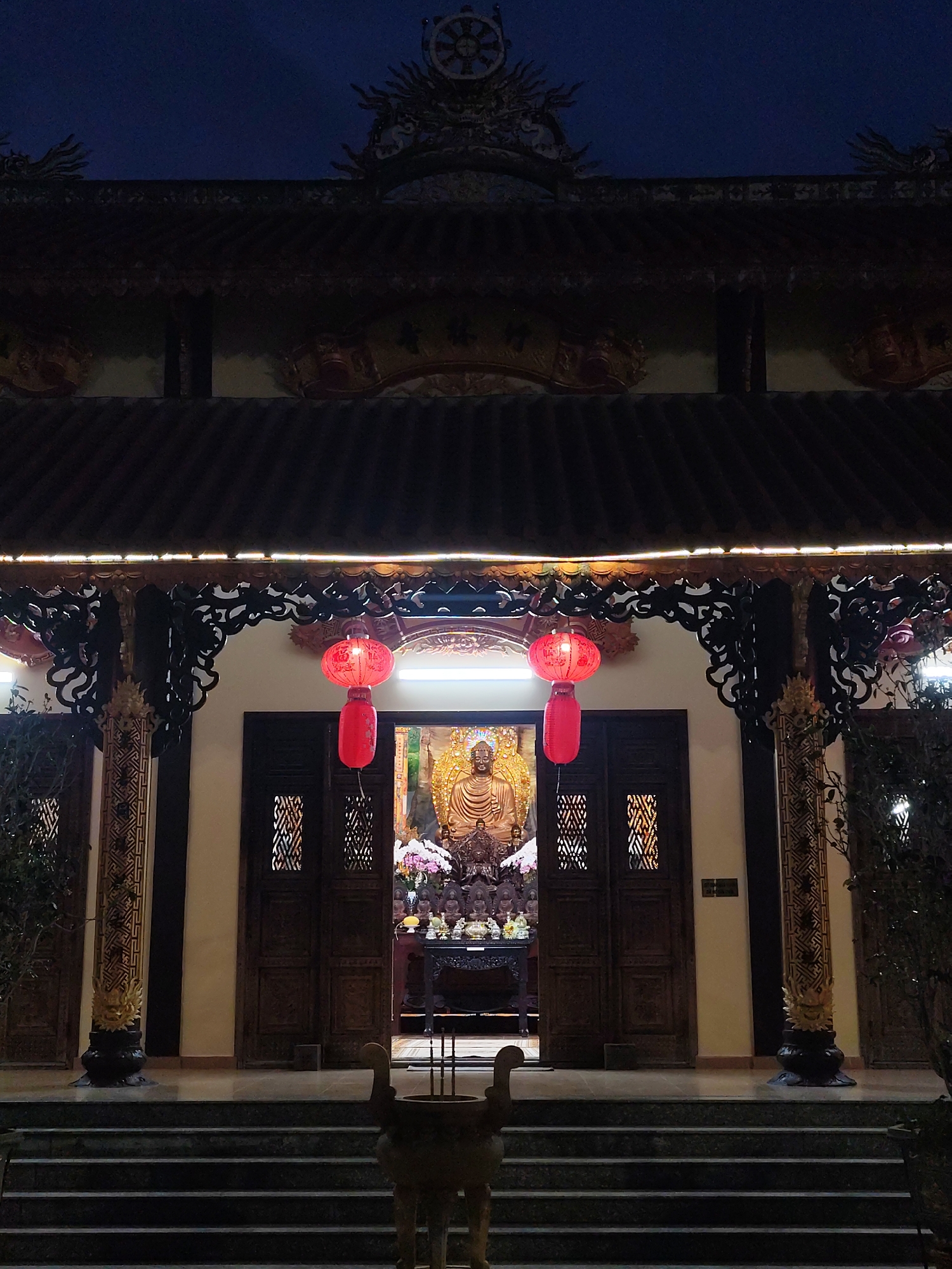 The image depicts the entrance to a Buddhist temple in Lâm Viên - Đà Lạt, Vietnam at night. The temple entrance features a multi-tiered roof with decorative trim and a Dharma wheel symbol at the apex. Two red lanterns hang on either side of the doorway. Inside, a large golden Buddha statue is visible, surrounded by offerings and other religious figures. The entrance is flanked by ornately carved pillars and wooden doors with lattice-like openings. Stone steps lead up to the entrance, and a bronze incense burner stands at the base of the stairs. The overall lighting is dim, with the interior of the temple and the lanterns providing the main sources of illumination.