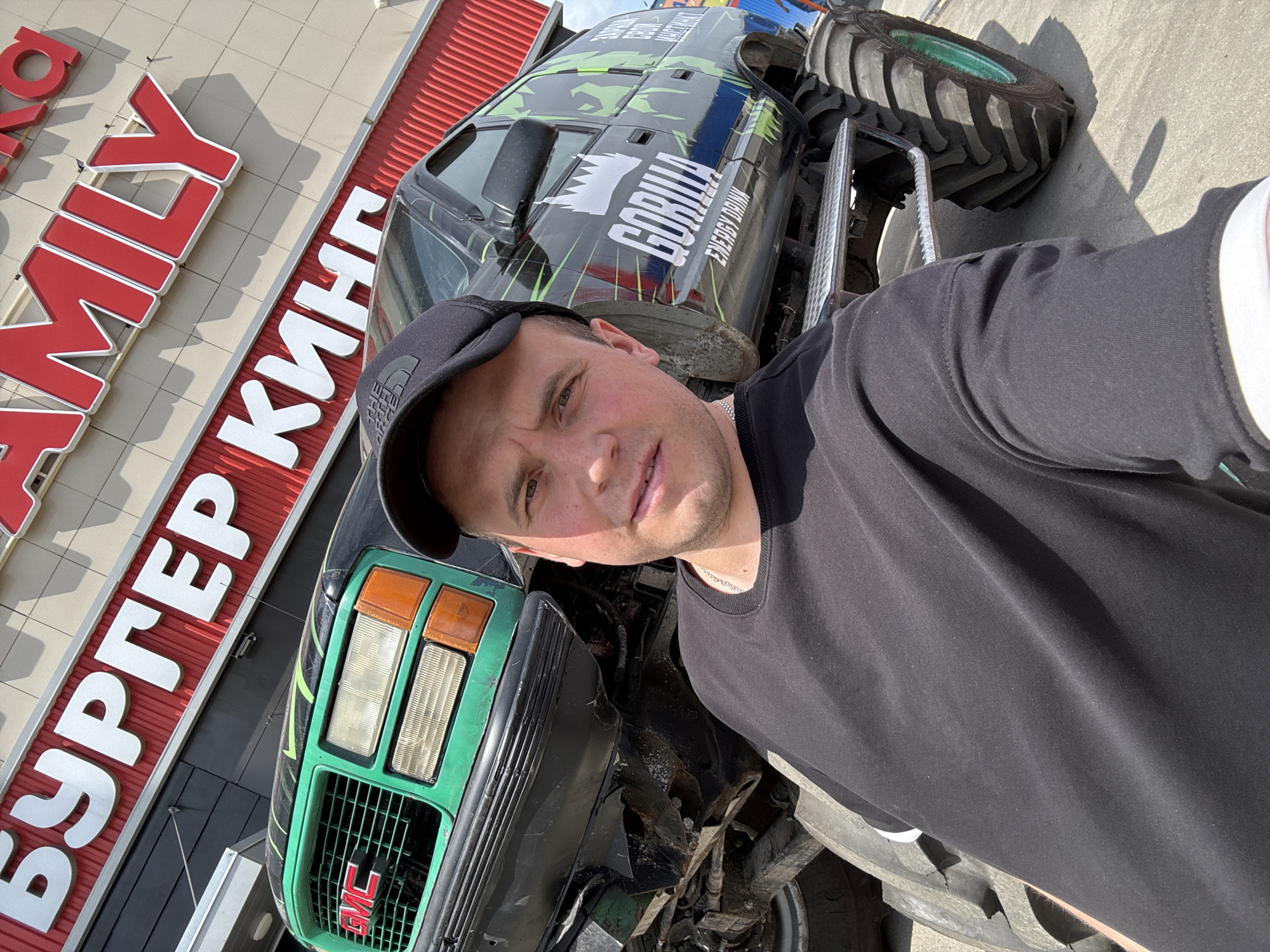 The image shows a man taking a selfie in front of a heavily modified GMC truck with oversized tires. The truck is decorated with "Gorilla Energy Drink" branding. Behind the man and the truck is a building with signage in Russian that translates to "Burger King Family." The location is Izhevsk, Russia. The man is wearing a black t-shirt and a black cap. The truck's front end, with its green grill and GMC logo, is prominently featured. The image appears to be taken during daylight hours.