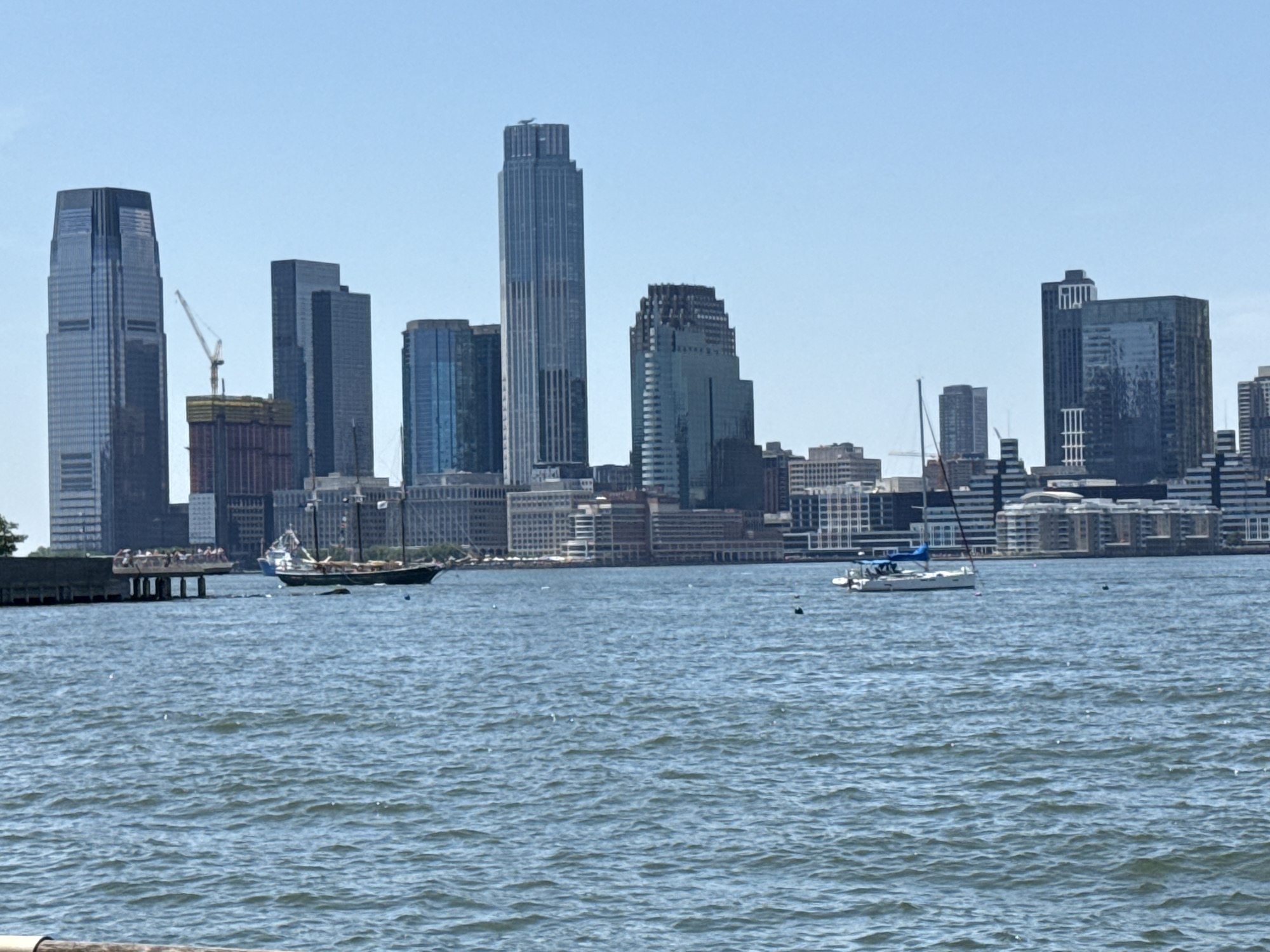 The image depicts a cityscape of skyscrapers and a body of water. In the foreground, the water is choppy with small waves, suggesting a breezy day. Two sailboats are visible on the water. One is a larger, older-fashioned sailing ship with multiple masts and sails, while the other is a smaller, modern catamaran with a blue canopy. In the middle ground, a pier or boardwalk extends into the water, with a few people standing on it. Beyond the water, a dense collection of skyscrapers of varying heights and architectural styles dominates the skyline. The buildings are modern, with glass facades reflecting the clear blue sky. The sky is a bright, cloudless blue. The scene appears to be a waterfront view of a major metropolitan area, likely New York City, given the iconic architecture. The lighting indicates it is daytime.