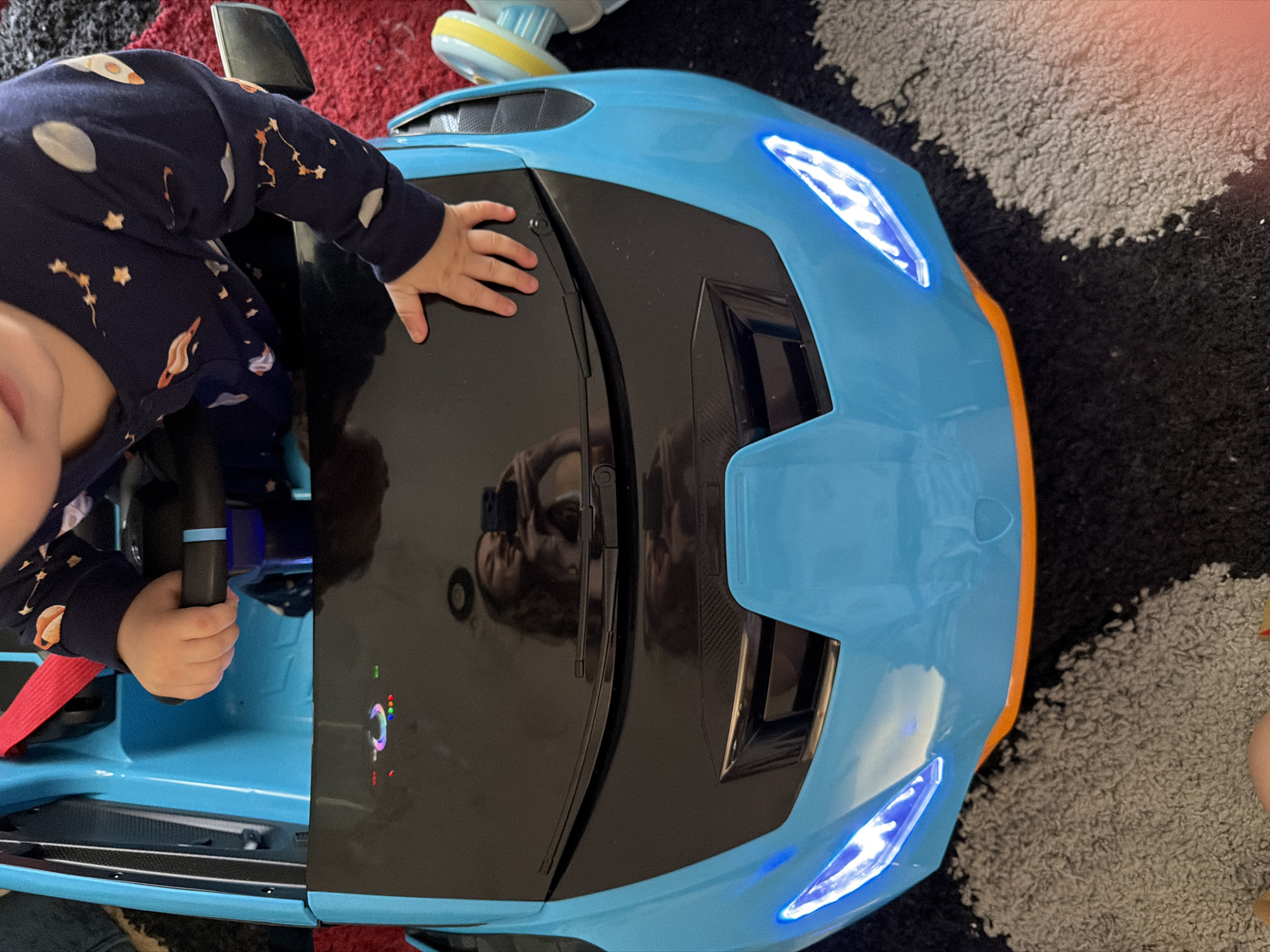 A toddler wearing pajamas with a celestial print is holding the steering wheel of a bright blue toy car. The car has a black hood with some illuminated buttons. The toddler's hand is visible gripping the black steering wheel, and another hand is resting on the hood. The headlights of the toy car are illuminated with a bright blue light. A reflection of a woman holding a phone, likely taking the picture, is visible on the car's hood. The car is positioned on a carpeted floor. The background shows a red rug and part of another toy. There is no indication of the city of Sharm El Sheikh, Egypt, in the image.