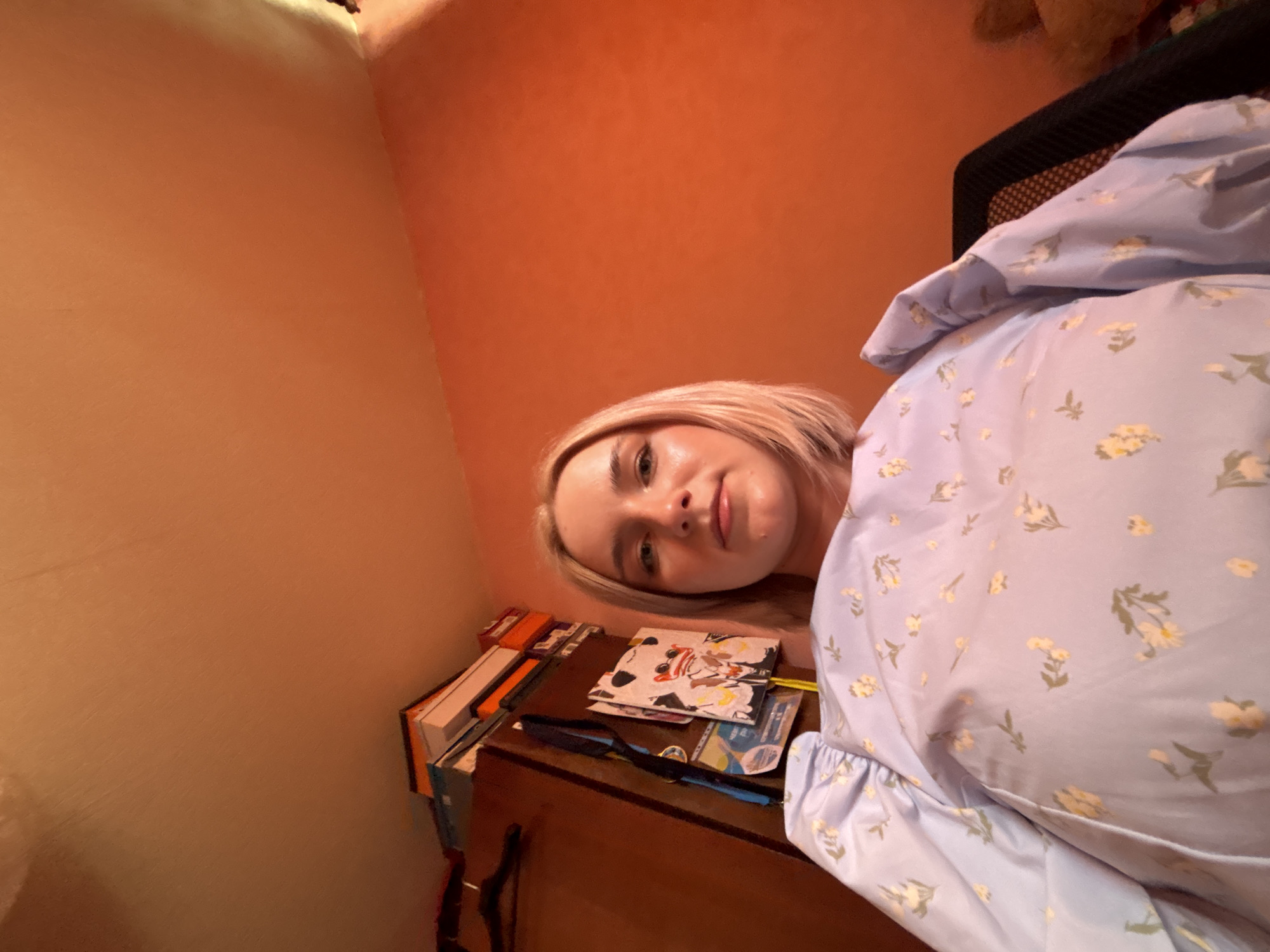 This low-angle portrait captures a young woman with blonde hair, fair skin, and light eyes, looking directly at the camera with a soft, slightly smiling expression. She is wearing a light blue garment, possibly a dress or blouse, patterned with small yellow and green flowers and leaves. The setting is indoors, with the background consisting of warm, uniform terracotta-orange walls.
Below her, a wooden table or desk is visible, holding a stack of books with orange and white spines on the left. Several flat items, including one with a stylized cat illustration, are also arranged on the table, alongside a black strap with blue accents. Partially visible behind her right shoulder is a dark-colored chair with a mesh back, and a blurry, dark brown, furry object is in the upper right corner of the frame. The lighting suggests an indoor environment, possibly illuminated by artificial light or warm ambient light. The image appears to be a casual selfie taken in a personal space, likely in Saint Petersburg, Russia. No discernible text is visible.
