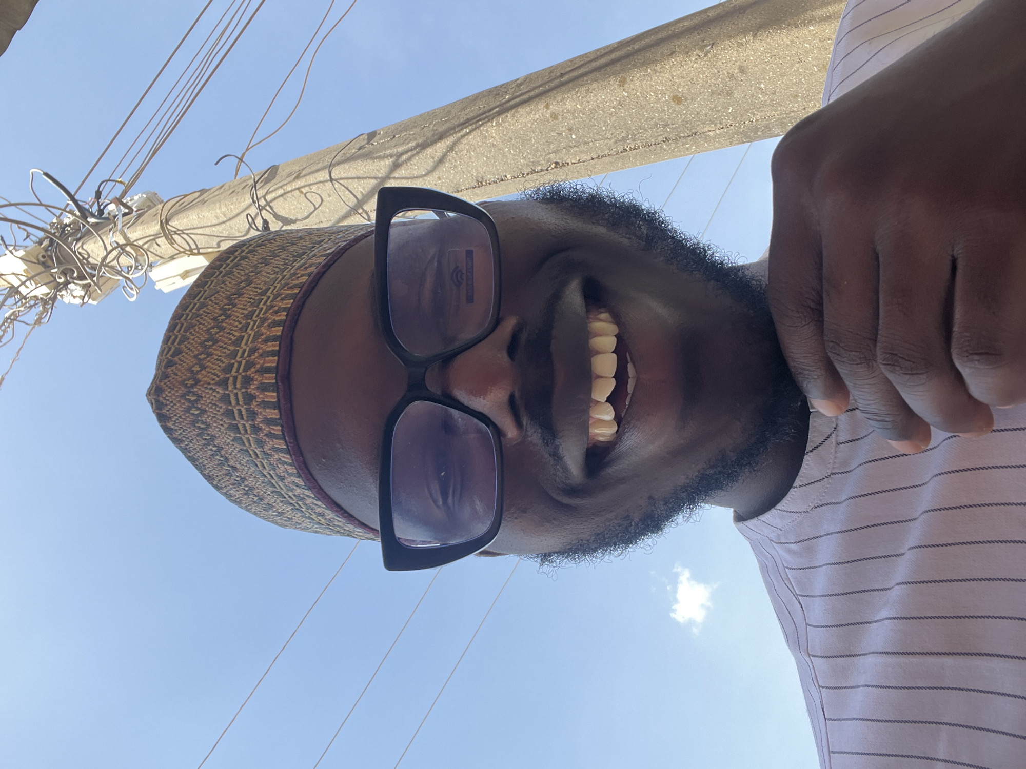 A selfie was taken outdoors by a Black man wearing glasses and a patterned cap. He is smiling broadly, showcasing his teeth. The image is taken from a low angle, capturing a utility pole, a web of wires, and a clear, blue sky in the background. The man is wearing a striped shirt. Given the context of Jos, Nigeria, the scene likely reflects an urban or suburban environment within the city.