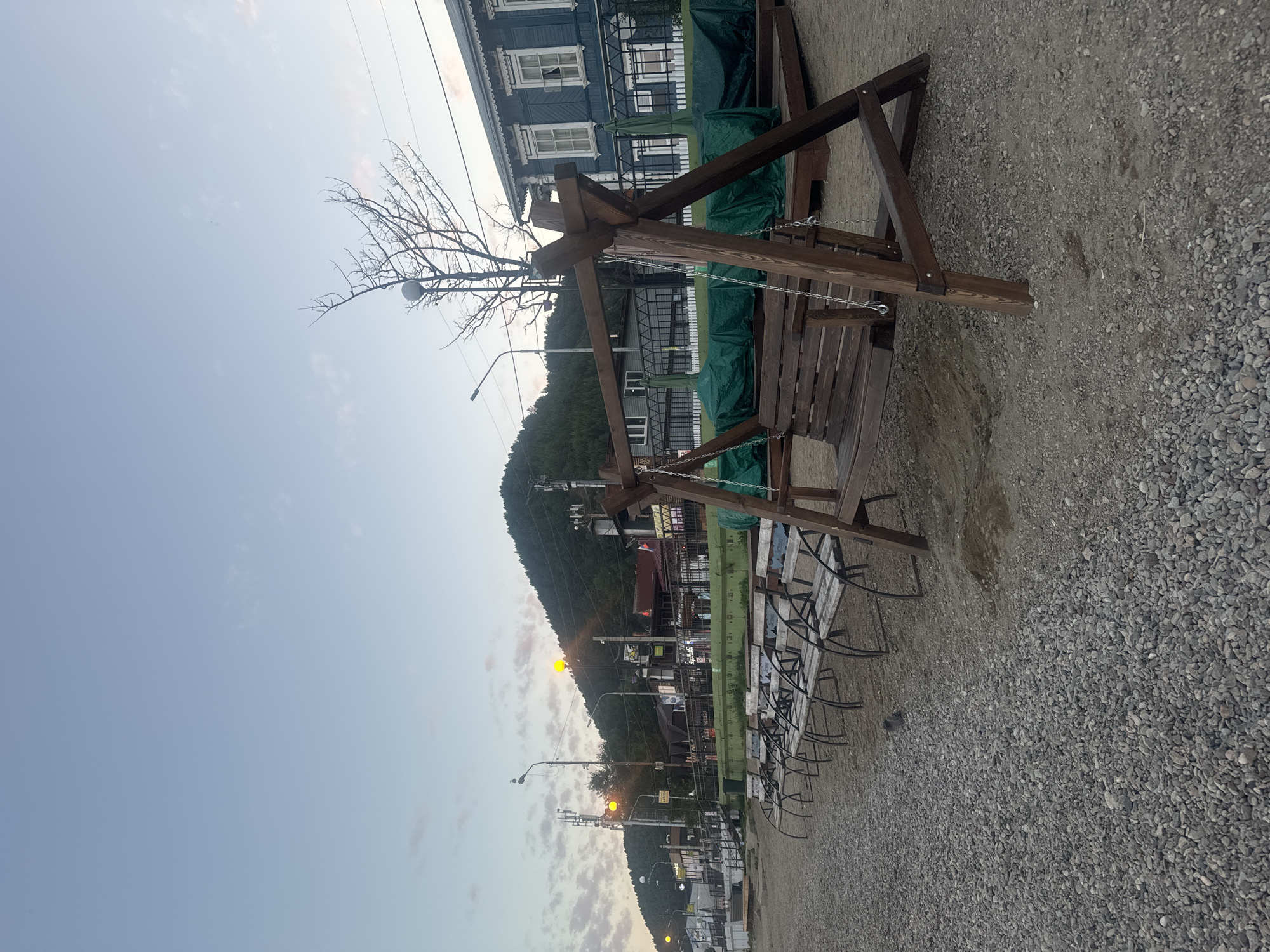 The image shows a public area in Listvyanka, Russia, along the shore of Lake Baikal, during twilight. In the foreground, a wooden swing set is prominently featured on a sandy and pebbled area. Behind the swing, a row of empty wooden benches with metal frames faces away from the viewer, suggesting a seating area for visitors. Further back, a row of green tarps covers outdoor seating or tables. Beyond this, a blue wooden building with white decorative trim and windows stands out. The background is dominated by a steep, forested hill, with buildings and streetlights visible along its base, indicating a town situated on the incline. The sky is a pale blue with some wispy clouds, and a few yellow lights are illuminated on street lamps and building facades, hinting at the approaching evening. A lone bird is visible on the ground in front of the benches. The overall scene depicts a quiet, recreational waterfront area in a lakeside town.