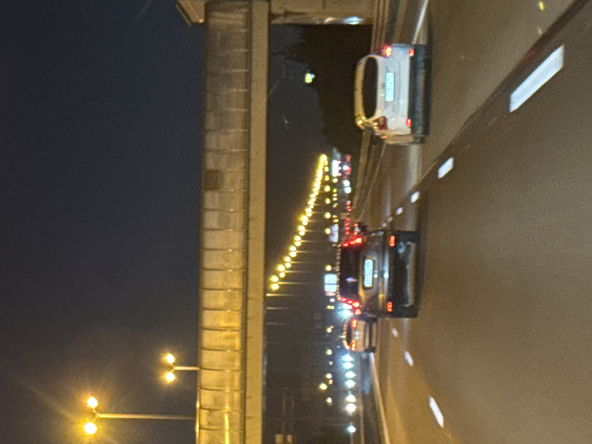 The image depicts a nighttime view of traffic on a highway in Saint Petersburg, Russia. A pedestrian overpass is visible spanning the highway, illuminated by yellow lights. Several cars are on the road, their headlights and taillights creating streaks of light. The scene is characterized by the dark sky and the artificial lighting from the overpass and streetlights. The overall impression is one of a typical urban highway at night.