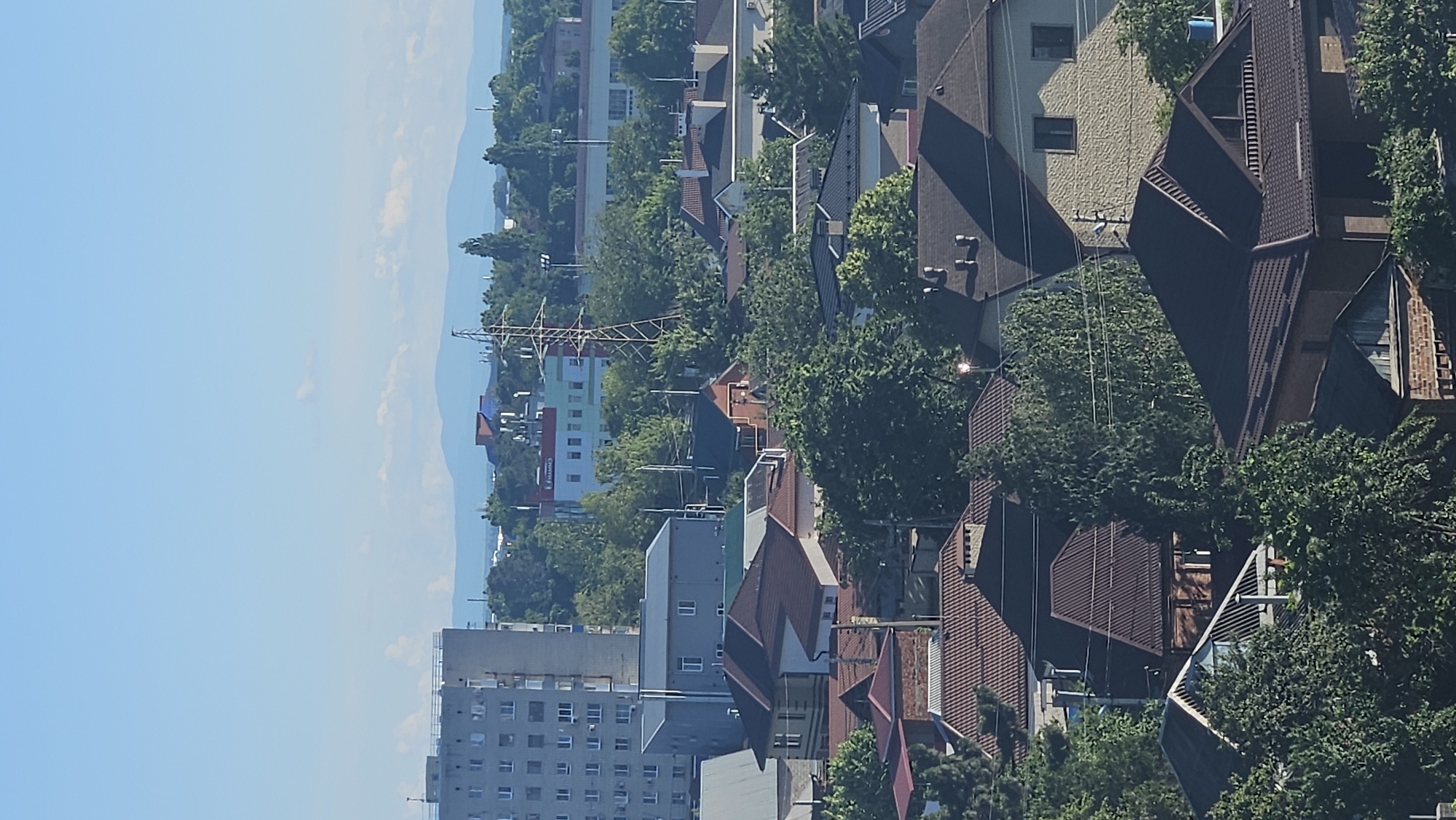 This is an aerial photograph of a residential area in the city of Krasnodar, Russia, under a clear blue sky. The scene is dominated by numerous houses with various roof styles and colors, primarily brown and red tiles. Interspersed among the houses are lush green trees, creating a verdant landscape. In the background, a tall, grey, multi-story building is visible, along with other urban structures and a prominent power line tower. Further in the distance, hazy blue mountains are discernible on the horizon. There are no people or vehicles depicted in the image. The overall impression is a sunny day in a densely populated suburban or semi-urban area.