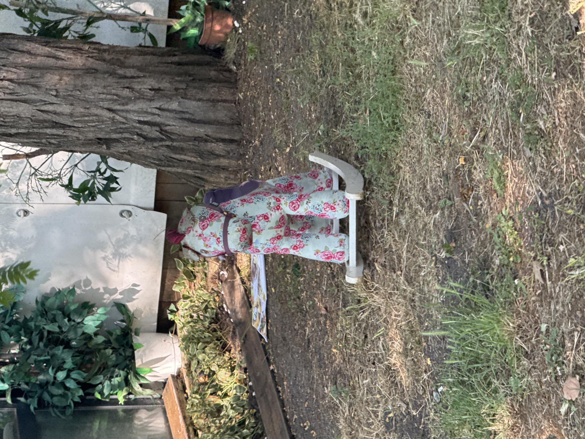 The image depicts a rocking horse toy with a floral pattern, positioned outdoors on a patch of dry grass and dirt. A large tree trunk is visible in the background, along with some greenery and a white structure. There are no people or any specific elements that would suggest a location in Burgas, Bulgaria. The scene appears to be a yard or garden area.