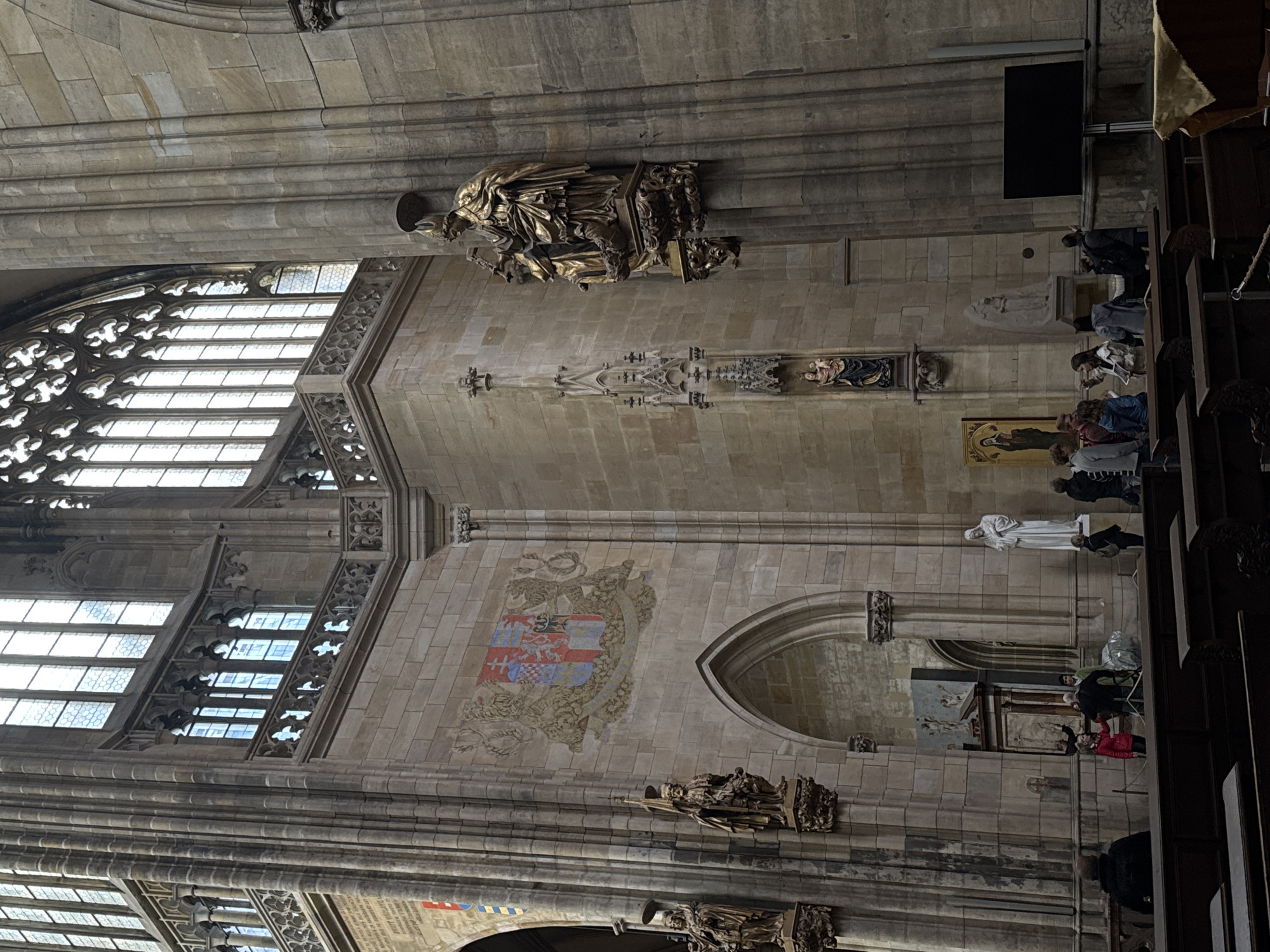 The image shows the interior of a large, stone cathedral. The architecture is Gothic, with high vaulted ceilings and large stained-glass windows. There are several statues and ornate carvings visible. In the foreground, there are rows of dark wooden pews.
In the middle ground, a group of people are gathered, appearing to be tourists. They are looking at various architectural features and artworks. Some are pointing and gesturing. One person in a red jacket is talking to another individual. There is a barrier cordoning off an area.
In the background, the grandeur of the cathedral continues, with more statues, a large crest displayed on the wall, and a tall, fluted column. The lighting suggests it is daytime, with light filtering through the windows. The overall atmosphere is one of reverence and historical significance.