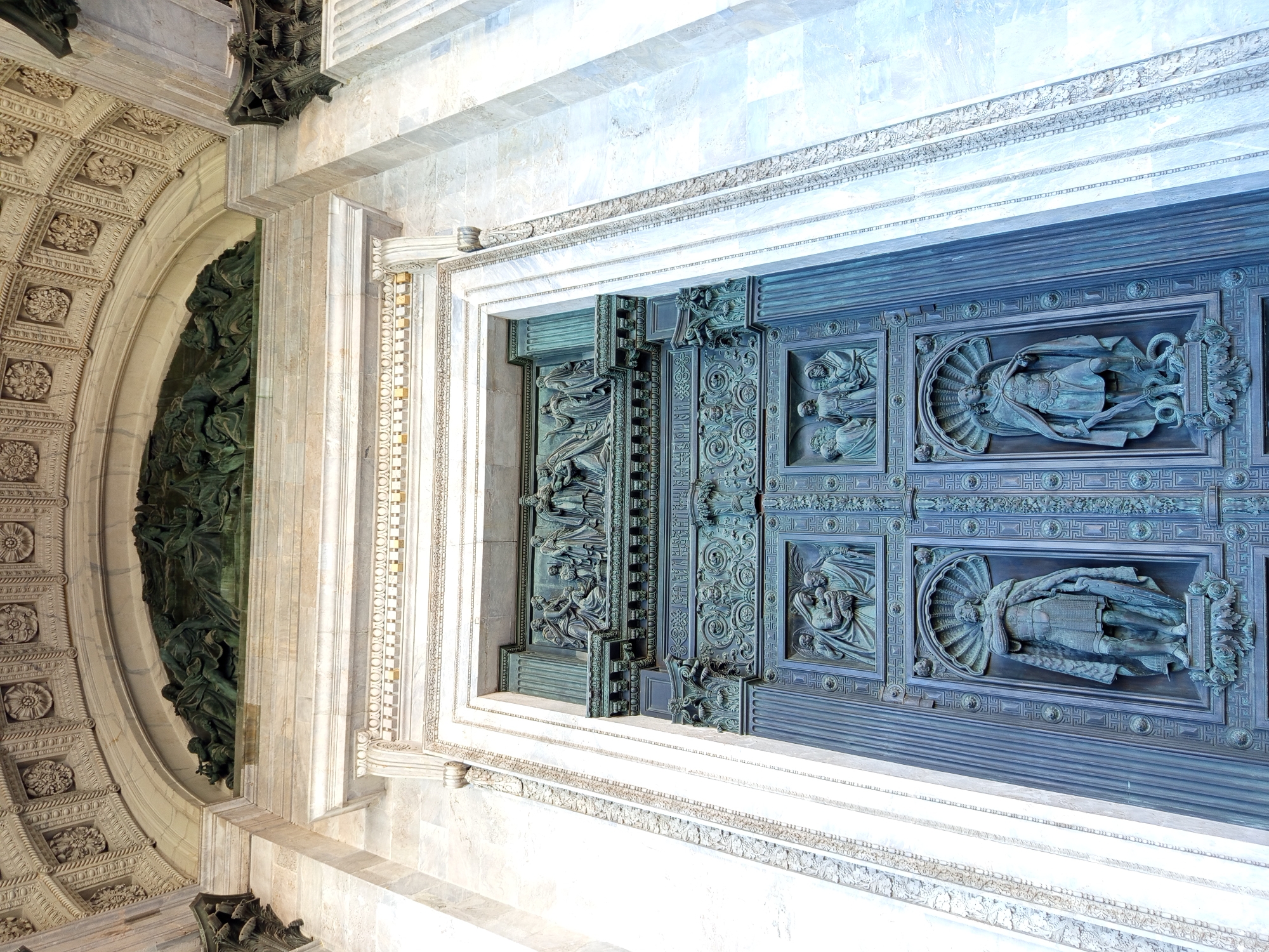 The image shows a close-up, low-angle view of a monumental bronze doorway and adjacent architectural elements, suggesting a grand, classical building. The doorway is comprised of large, intricately carved panels, each depicting figures in relief. The style of these figures suggests a religious or historical theme, with draped robes and dramatic poses.
The doorway is framed by ornate stonework, including columns, cornices, and friezes. The stonework itself is highly detailed, featuring repetitive patterns and sculptural embellishments. To the left of the doorway, a large, arched opening reveals a dark, green-hued sculptural relief, appearing dense with many figures. The overall impression is one of immense scale and meticulous craftsmanship.
The lighting suggests it is daytime, with bright illumination highlighting the textures of the stone and metal. The blue-green patina on the bronze doors indicates their age and exposure to the elements. The scene is static, focusing on the architectural and sculptural details rather than any human activity. The location is not explicitly identifiable, but the style of architecture suggests a European setting, possibly a cathedral or a significant public building. No visible text is present in the image.