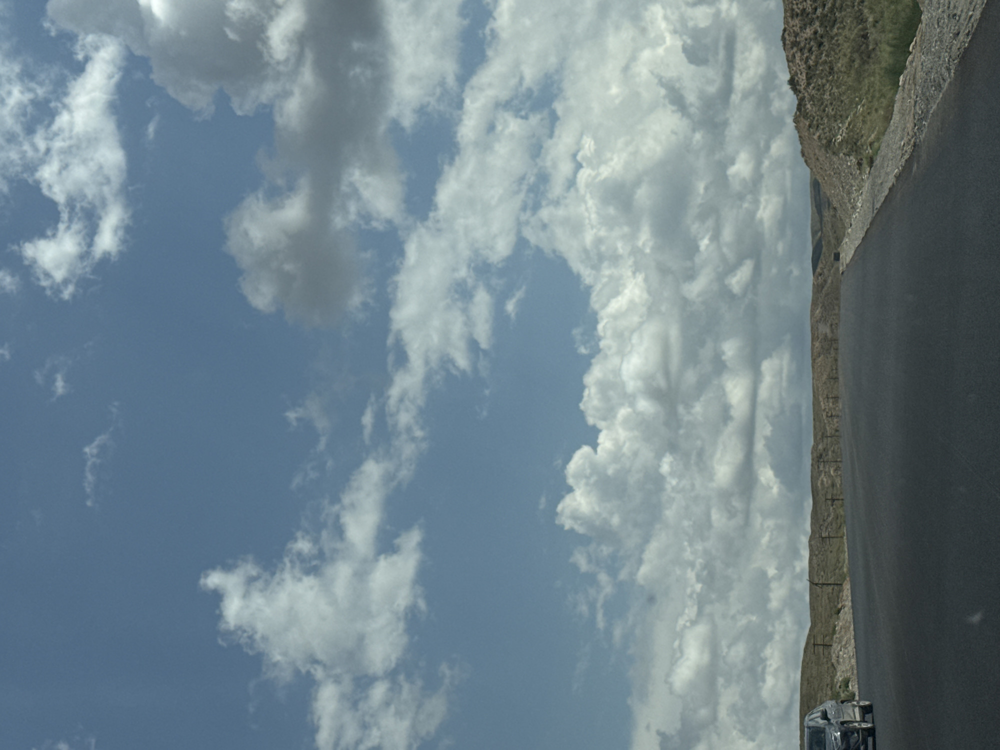 The image captures a scenic view of a road curving through a landscape in the city of Kyzyl-Tuu, Kyrgyzstan. The sky is a vibrant blue, adorned with fluffy white clouds. On the left side of the road, a dark-colored SUV is partially visible, traveling in the same direction as the camera. The road itself is paved and appears to be in good condition, with a slightly rough texture. To the right of the road, the terrain rises into rolling hills covered in dry grass and sparse vegetation. A simple wire fence runs along the edge of the road, separating it from the hilly landscape. In the distance, the hills continue to undulate, disappearing into a hazy horizon. The overall impression is one of a peaceful, rural setting under a dynamic sky.