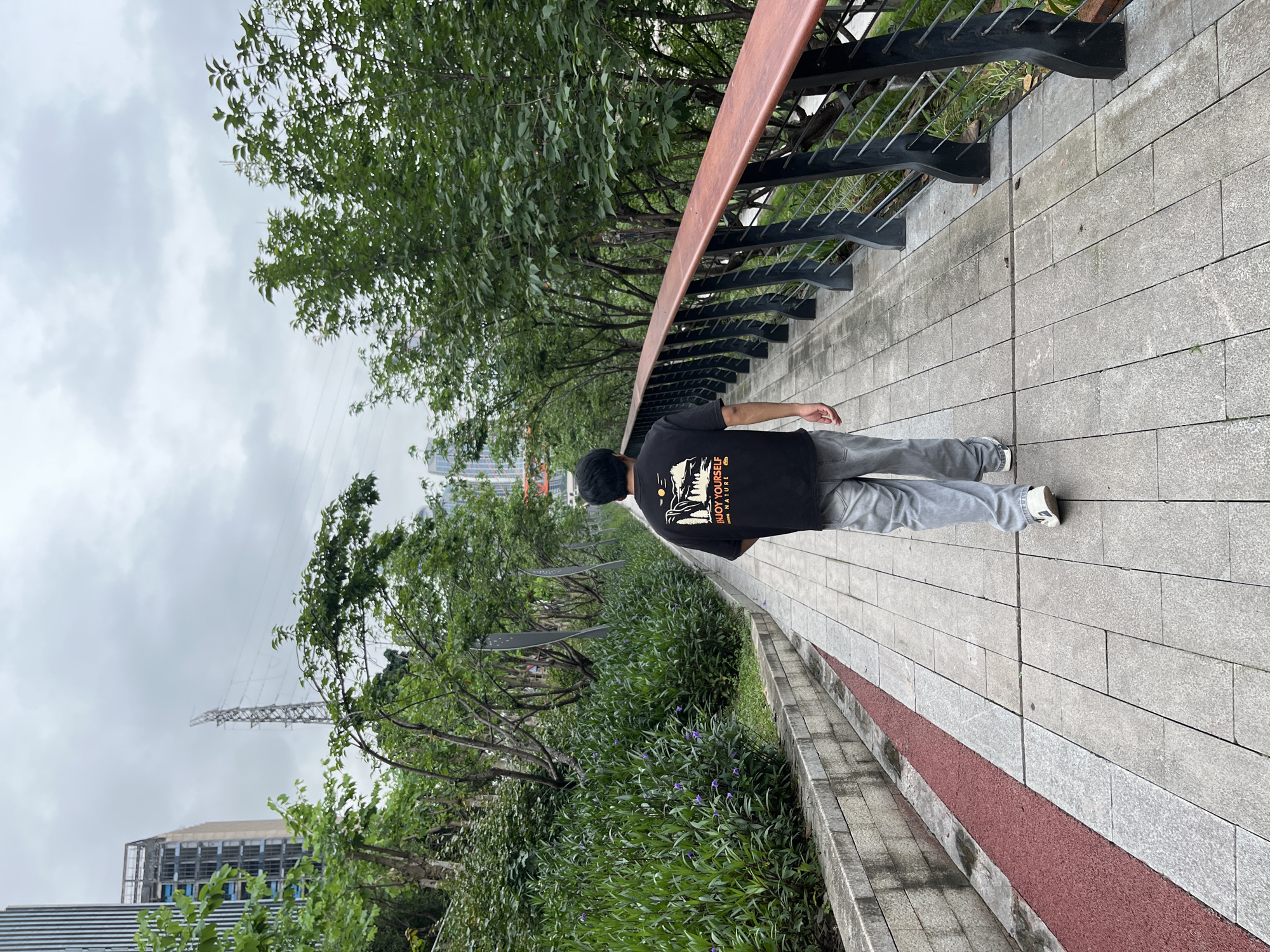 A man wearing a black T-shirt and gray pants is walking on a paved walkway in Jiangbeicheng Subdistrict, China. The walkway is lined with trees and vegetation on one side and a metal railing with a wooden top on the other. The sky is overcast. In the distance, buildings and a power pylon are visible. The walkway also has a red section.