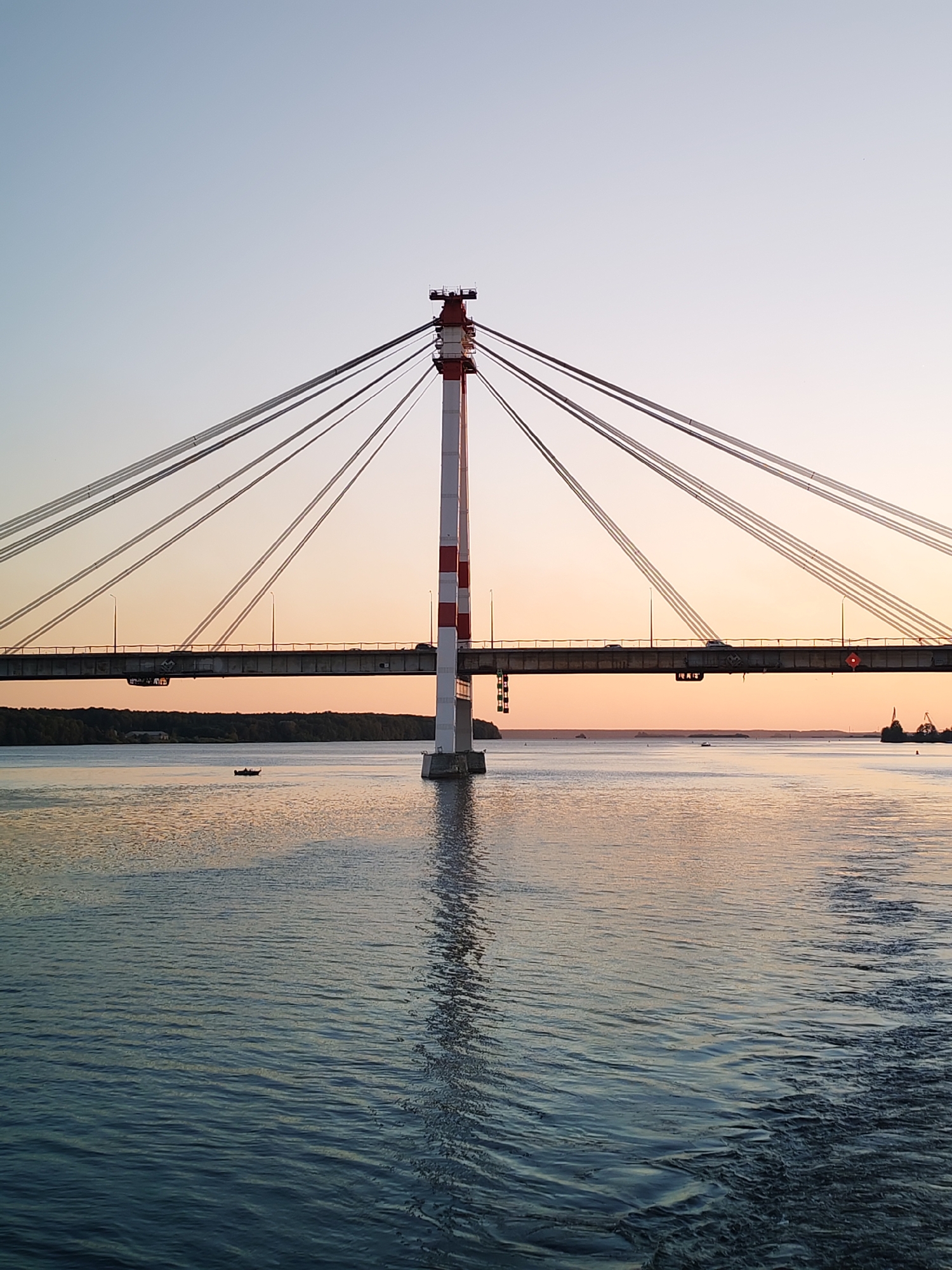 This is a photograph of the Yagorbsky Bridge in Cherepovets, Russia, taken at sunset. The bridge is a cable-stayed bridge with a distinctive red and white striped pylon in the foreground, supporting the bridge's structure with numerous cables. The roadway of the bridge is visible, with cars driving across it. The bridge spans a wide body of water, likely the Rybinsk Reservoir, which is calm and reflects the warm hues of the sunset. In the distance, a tree-lined shore is visible. A small boat with two occupants is positioned on the water in the middle ground. The sky transitions from a pale blue at the top to a soft orange and pink gradient as it nears the horizon, indicating the time of day is sunset. The overall scene is serene and captures the bridge and its surroundings in a soft, ambient light.