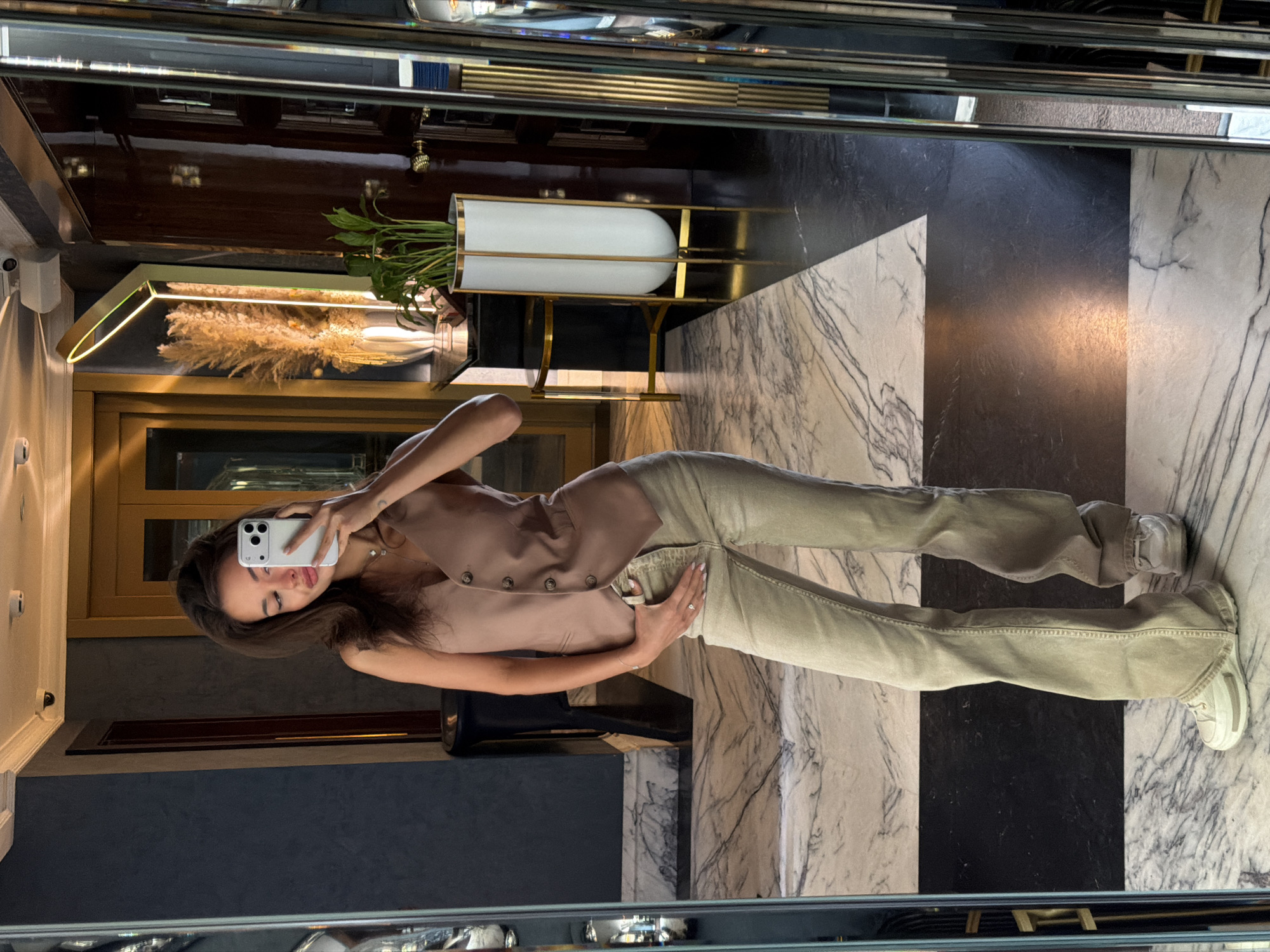 A young woman is captured in a full-length mirror selfie within an upscale, modern interior, likely a hotel lobby or a luxurious building in Moscow, Russia. She has long dark hair and is holding a white smartphone to take the picture, her gaze directed towards the phone screen. She is wearing a light brown or beige sleeveless vest over a lighter top, paired with light green wide-leg pants and white sneakers. Her right hand is casually placed near her hip.
The background, reflected in the mirror, features refined decor. To her right, a tall, white cylindrical planter holding green leafy plants is set on a minimalist gold metal stand. Below it, a rectangular display box with a gold frame contains dried pampas grass or similar decorative reeds, illuminated by a strip light. Dark wooden paneling or a door is visible further back. The ceiling above appears to be mirrored as well, reflecting portions of the room.
The floor is a striking element, composed of large rectangular tiles arranged in an alternating pattern of white marble with distinctive black veining and solid black marble. The overall ambiance is luxurious and sophisticated, characterized by polished surfaces, metallic accents, and elegant design. No visible text is present in the image. The time of day is not discernible due to the indoor setting and artificial lighting.