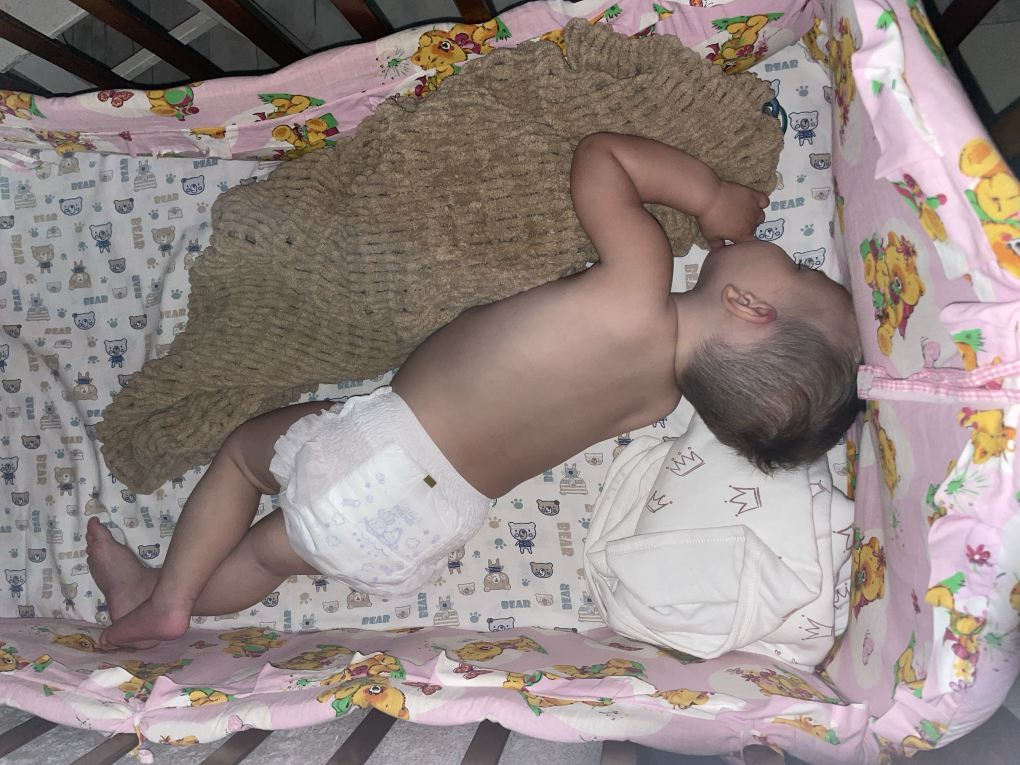 A baby is asleep in a crib. The baby is lying on their side, wearing a diaper, and partially covered by a brown, furry blanket. The crib has patterned sheets with bears on them, and a padded bumper around the edge with illustrations of Winnie the Pooh characters. The location is unspecified, and there is no indication it is in Perm, Russia.