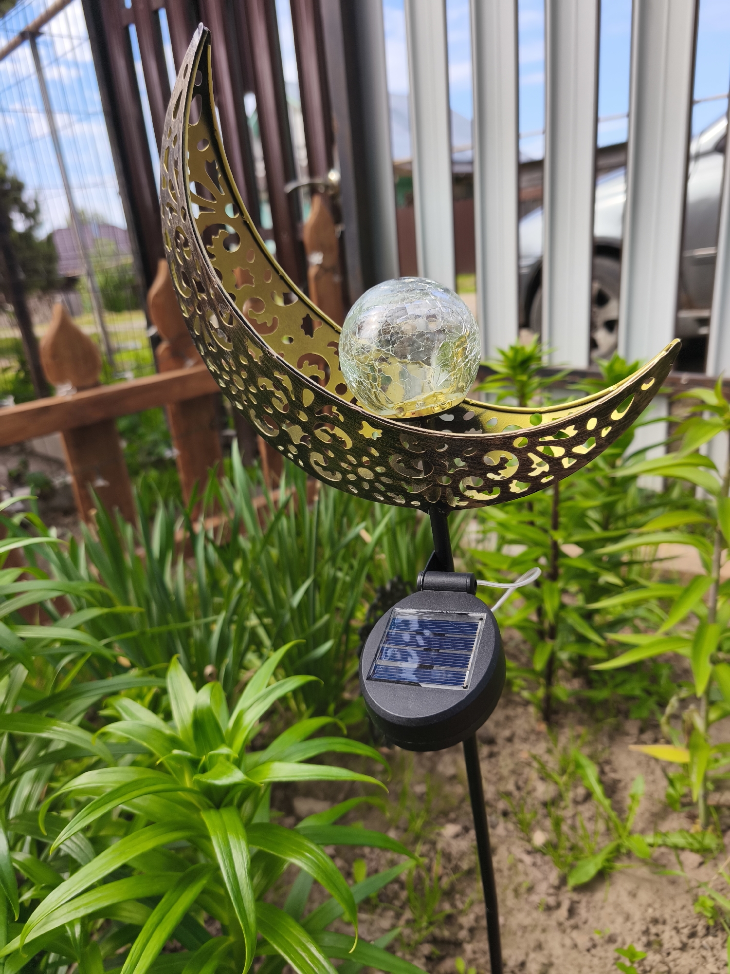 A close-up, eye-level shot features a solar-powered garden light in the shape of a crescent moon with a crackled glass ball at its center. The moon is a metallic gold color and has intricate cut-out patterns. Beneath the moon, a black solar panel on a stake is visible. The garden light is positioned amongst lush green plants with pointed leaves. In the background, a brown wooden fence separates the garden from a light-colored, vertical-slatted fence behind which a car is partially visible. The sky above is blue with scattered white clouds. The scene appears to be a residential garden in a city, possibly Puchezh, Russia, given the overall aesthetic and the mention of Russian location. There are no people or animals present in the image. The lighting suggests it is daytime, with sunlight illuminating the scene.