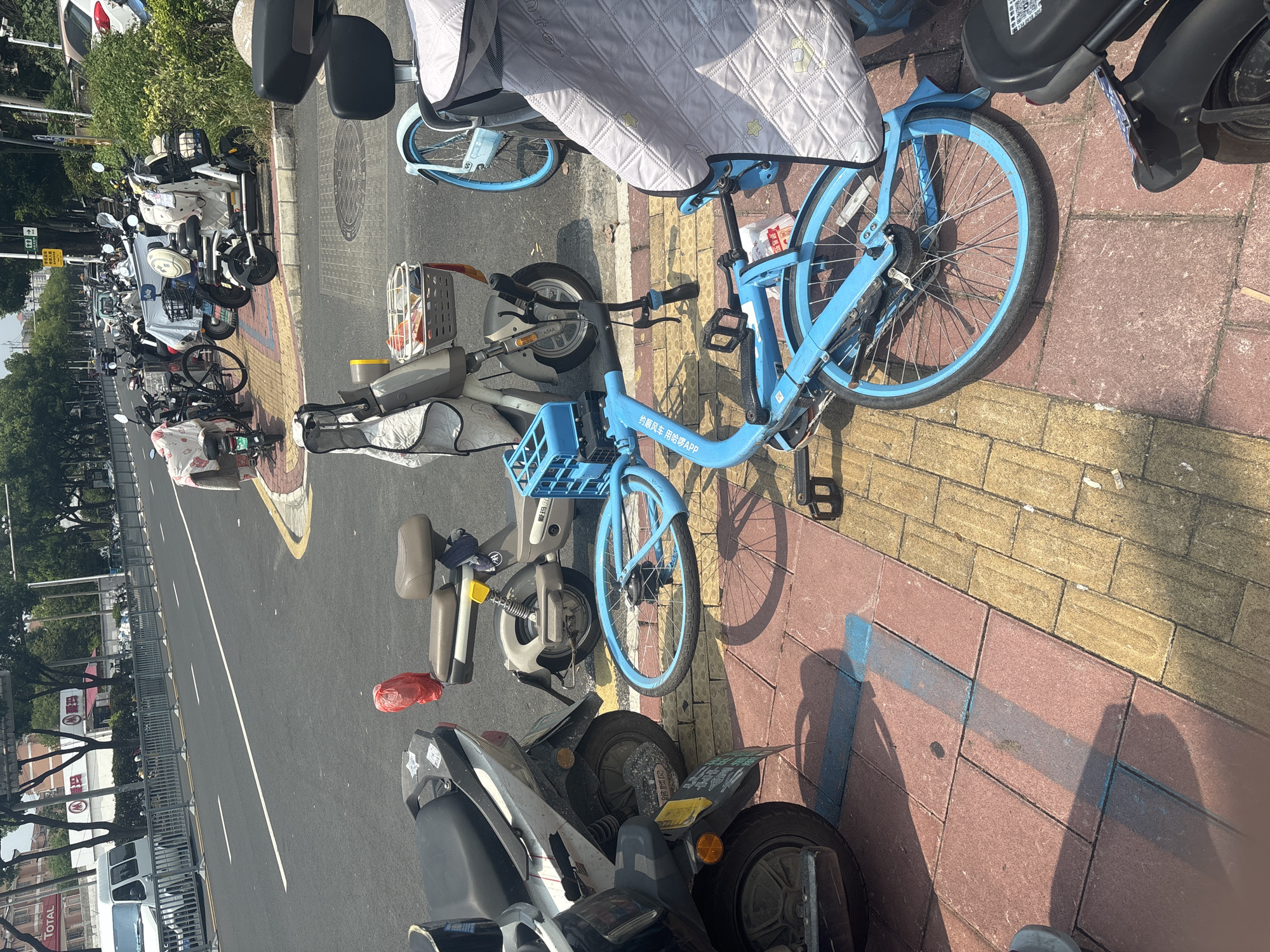 The image depicts a sunny outdoor scene in Suzhou Industrial Park, China, characterized by a street and sidewalk lined with various parked vehicles. In the foreground, a bright blue bicycle lies on its side on a paved walkway composed of red and yellow rectangular tiles. The bicycle's frame and wheels are prominently blue, and it features a basket on the front. Adjacent to the fallen bicycle are several parked electric scooters and motorcycles, some covered with light-colored cloths. One scooter, to the right of the blue bicycle, is also light-colored with a brown seat and a red plastic bag hanging from its handlebars. Further down the street, more scooters and bicycles are visible, parked along the roadside. Across the street, a white van is parked near a row of trees and buildings. The overall environment suggests a typical urban setting with available transportation options.