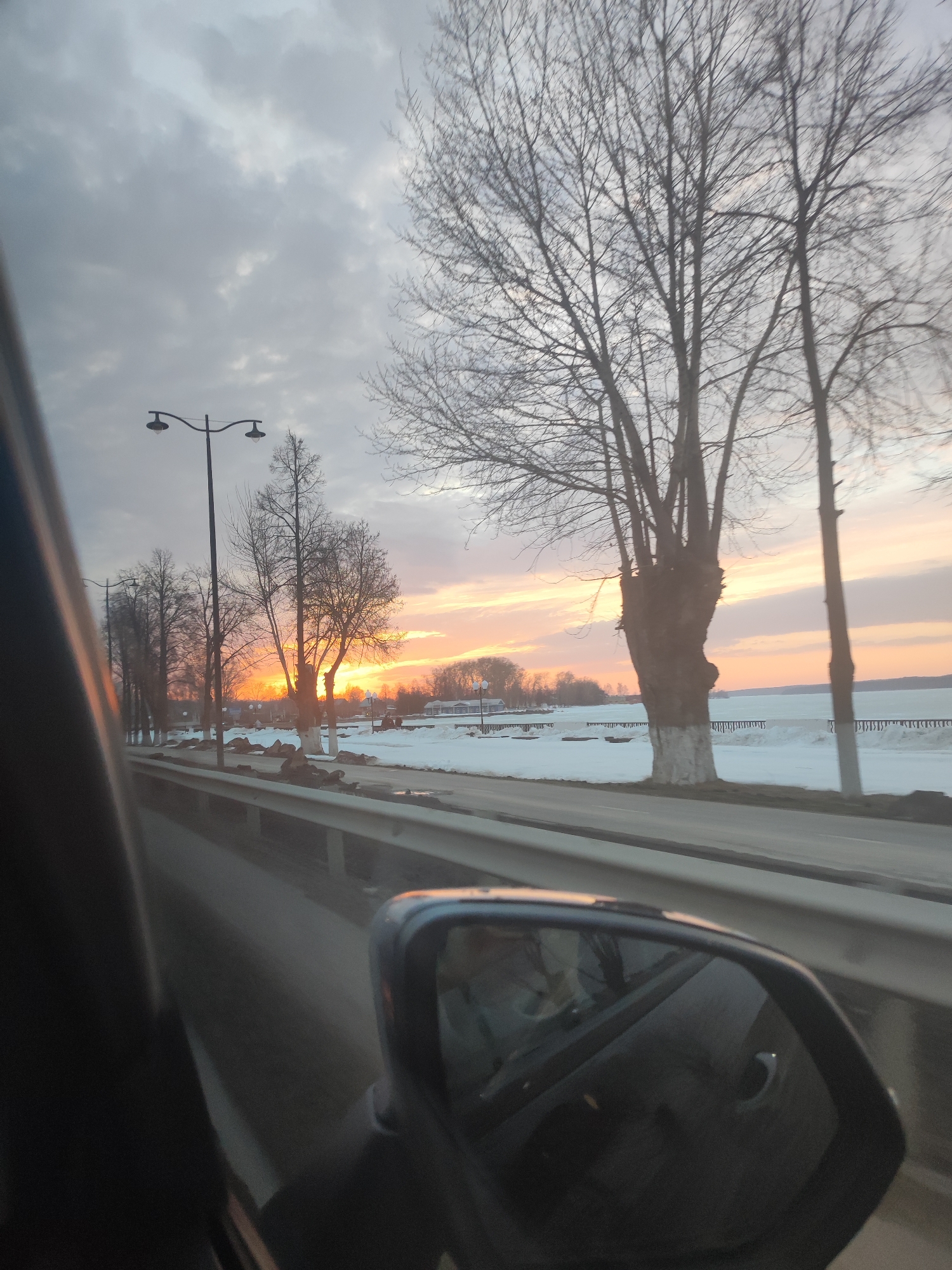 The image, taken from inside a car, shows a sunset over a snow-covered landscape in Votkinsk, Russia. The view is framed by the car's interior, including a side mirror. Bare trees line the road, some with whitewashed trunks. Streetlights are visible. A body of water with snow-covered shoreline is in the distance. The sky is a mix of gray clouds and orange hues from the setting sun.
