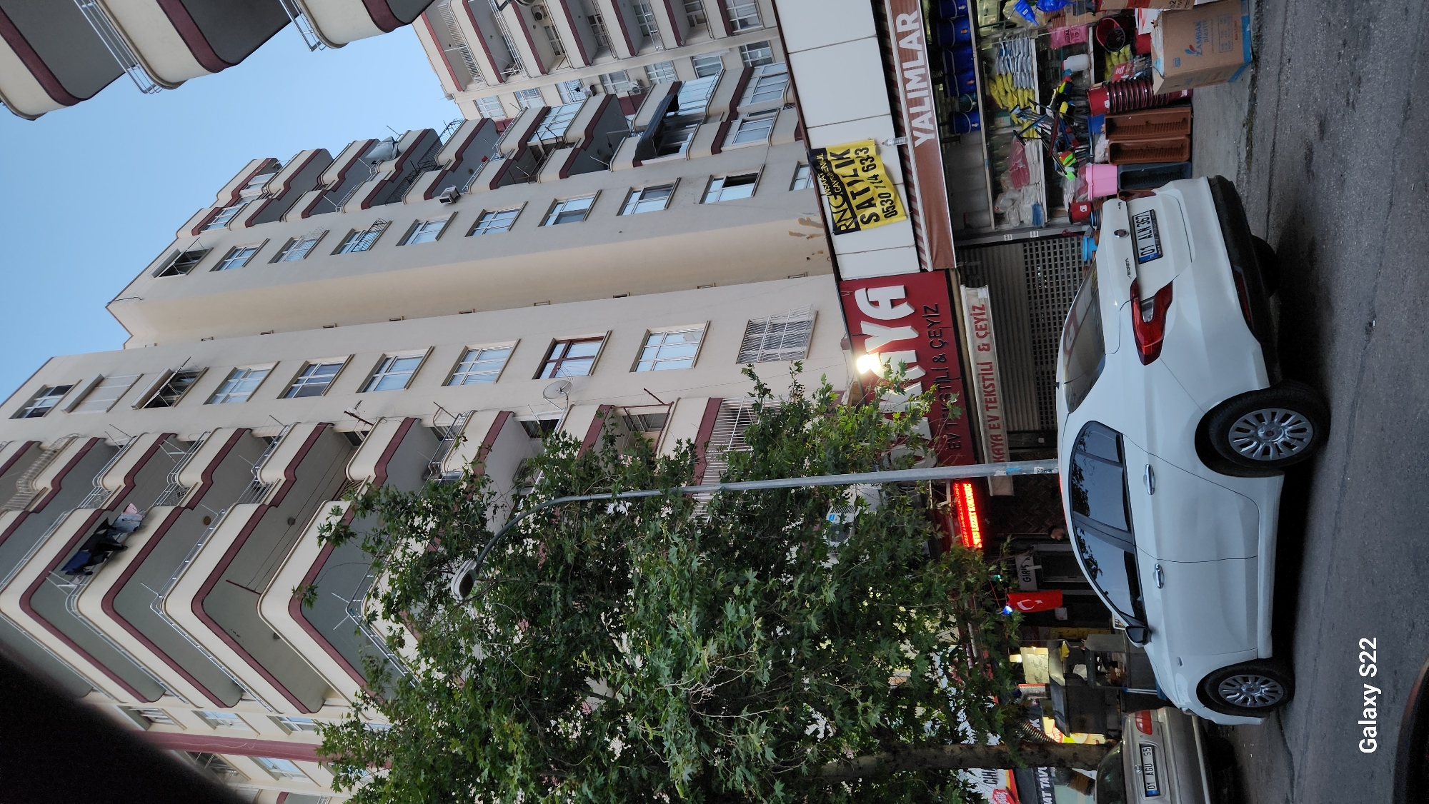 The image shows a street scene in Çukurova, Turkey, with a modern apartment building as the central focus. The building is tall and cream-colored with multiple balconies, many of which have dark red trim and metal railings. Clothes are hanging on a clothesline on one of the upper balconies. A large, leafy green tree is situated in the foreground on the left, partially obscuring the lower floors of the building. A street lamp is visible next to the tree. Below the apartments, a row of storefronts lines the street. One prominent storefront has a red and white sign that reads "KAYA EV TEKSTİLİ & ÇEYİZ." To the right of this, a yellow sign with black text says "SATILIK" (for sale) and a phone number. Further to the right, another storefront displays various products, including buckets and containers, with a sign that appears to read "YALIMLAR." A white sedan is parked on the street in front of the storefronts. The sky above is a clear, pale blue, suggesting it is either early morning or late afternoon.