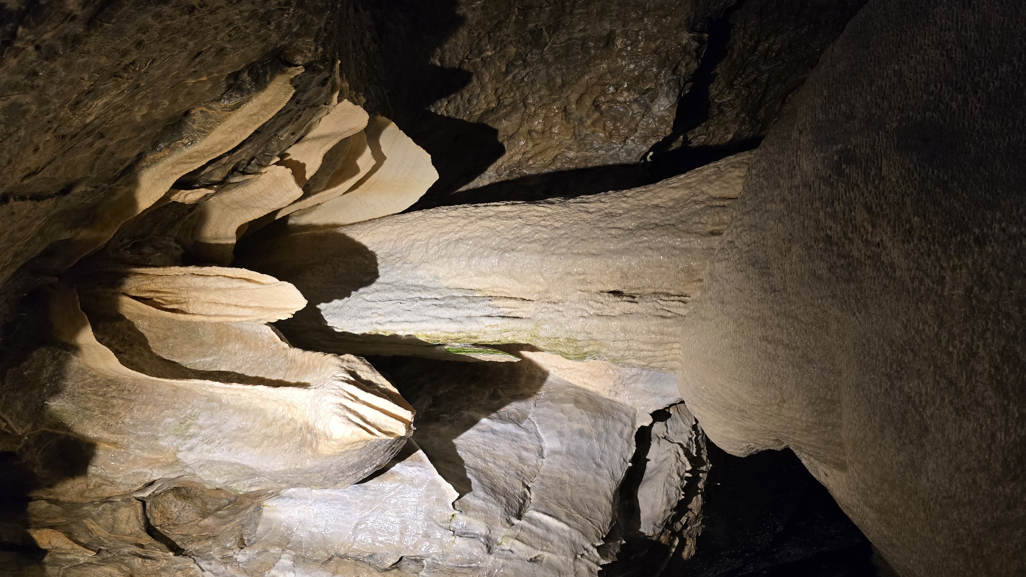 The image depicts the interior of a cave, likely a tourist attraction given the lighting and the clear formations. The primary subjects are the natural geological formations of stalactites and stalagmites, characterized by their layered, undulating textures and pale, earthy colors. These formations appear wet, with a sheen on their surfaces, suggesting the presence of water and active mineral deposition. The cave's ceiling and walls are rough and dark, contrasting with the smoother, lighter speleothems.
There is no activity or event taking place; the scene is static and captures a moment of geological time. Notable details include the intricate details of the rock formations, such as the rippled surfaces and sharp edges of the layered deposits, and the play of light and shadow, which accentuates the three-dimensional qualities of the cave's interior. The overall atmosphere is one of natural wonder and subterranean exploration. The lighting appears to be artificial, likely installed for visitor safety and illumination.
No people are visible in the image. No text is present. The location context of Vavřinec, Czechia, suggests this is a natural cave formation in that region. The time of day and weather are not determinable from the image, as it is an enclosed underground environment. The emotions evoked are likely awe and curiosity about the natural processes that create such formations.