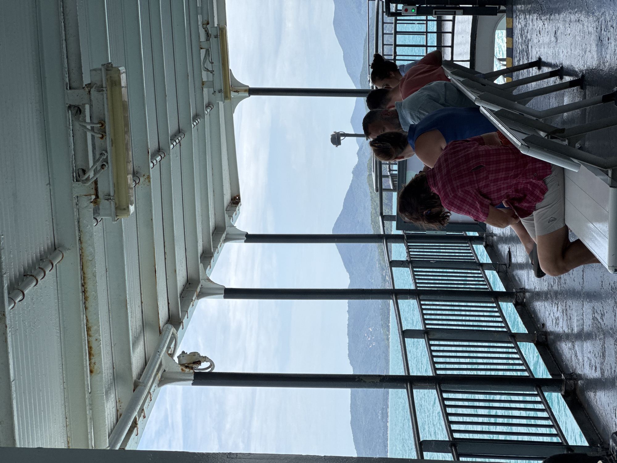 On a ferry crossing to Ko Pha-ngan, Thailand, passengers sit in rows of white plastic chairs, facing the turquoise sea. The ferry's overhead structure, a weathered white metal, shows signs of rust and is supported by black metal pillars. Beyond a black metal railing, lush green mountains rise from the water under a cloudy sky. The deck is wet and reflective, suggesting recent rain. The people are casually dressed, some looking out at the scenery, others conversing. The scene conveys a sense of peaceful travel and anticipation of arrival.