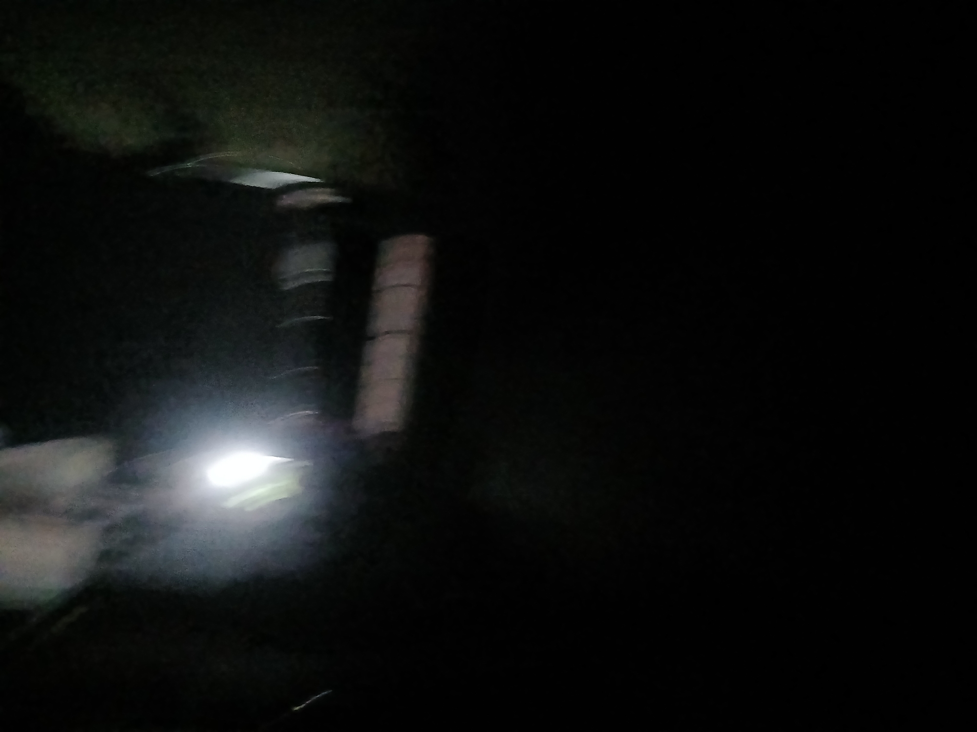 The image is a dark, blurry photograph taken at night. A bright light source, possibly a street light or headlights, is visible in the upper left quadrant, illuminating a misty area. Below the light, there is a dark horizontal structure with lighter, rectangular shapes, possibly part of a building or an object. The rest of the image is predominantly black, indicating a lack of ambient light. No specific features of Trușeni, Moldova, are discernible due to the poor quality of the photograph and the dark environment.