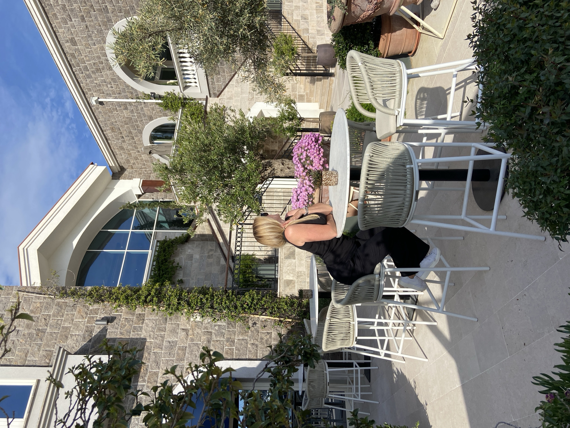 A bright outdoor scene captures a woman seated at a high-top table on a patio. The woman, with blonde hair, is wearing a black top or dress and white sneakers. She is positioned with her back partially to the viewer, looking slightly to her left.
The round, white table in front of her holds a potted arrangement of vibrant purple and pink flowers. Surrounding the table are three modern bar stools, featuring white frames and light grey woven seats. The patio itself is paved with light-colored stones, with various potted plants and shrubs scattered around, some casting distinct shadows on the ground due to the strong sunlight.
In the background, a large, multi-story building constructed from grey stone blocks dominates the scene. It exhibits varied architectural elements, including arched windows with white balustrades on an upper level and a striking large arched window with a white frame on a lower section. Green vines climb parts of the stone facade. Several olive trees with silver-green foliage are interspersed throughout the patio area, providing natural shade and a Mediterranean ambiance. Beyond the immediate seating area, more stone steps and additional tables and chairs are visible, suggesting a larger outdoor dining or lounging space.
The clear blue sky with a few wispy clouds indicates excellent weather. The strong sunlight and well-defined shadows suggest it is mid-morning or early afternoon. The overall setting evokes a relaxed and upscale outdoor environment, consistent with a villa or resort in a sunny location like Radovići, Montenegro. No visible text is present in the image.