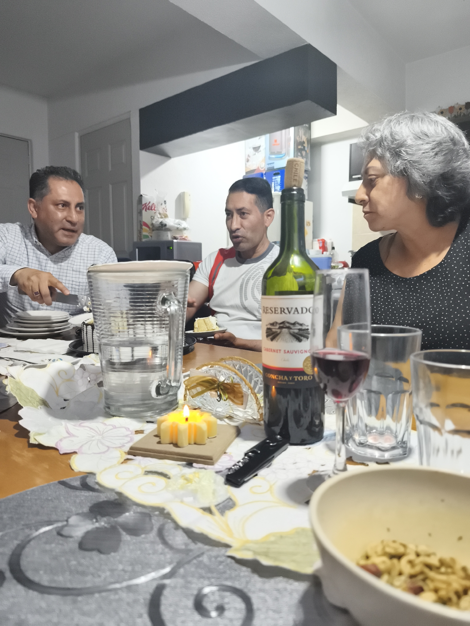 Three adults are gathered around a dining table in an indoor setting. One man, wearing a light-colored plaid shirt, is on the left side of the frame and appears to be cutting a slice of cake. Another man, wearing a white t-shirt with red accents, is seated to the right of the first, holding a plate with a slice of cake. A woman with gray hair, wearing a black and white polka dot shirt, is seated on the right side of the frame, looking towards the two men.
The table is covered with a patterned cloth in shades of gray, white, and yellow. There is a water pitcher, a bottle of wine with a cork, a wine glass filled with red wine, drinking glasses, and a lit candle on the table. A bowl filled with food is in the foreground.
In the background, there is a doorway, a dark-colored shelf, and some items on a shelf. The location is suggested to be Mexico City, Mexico.