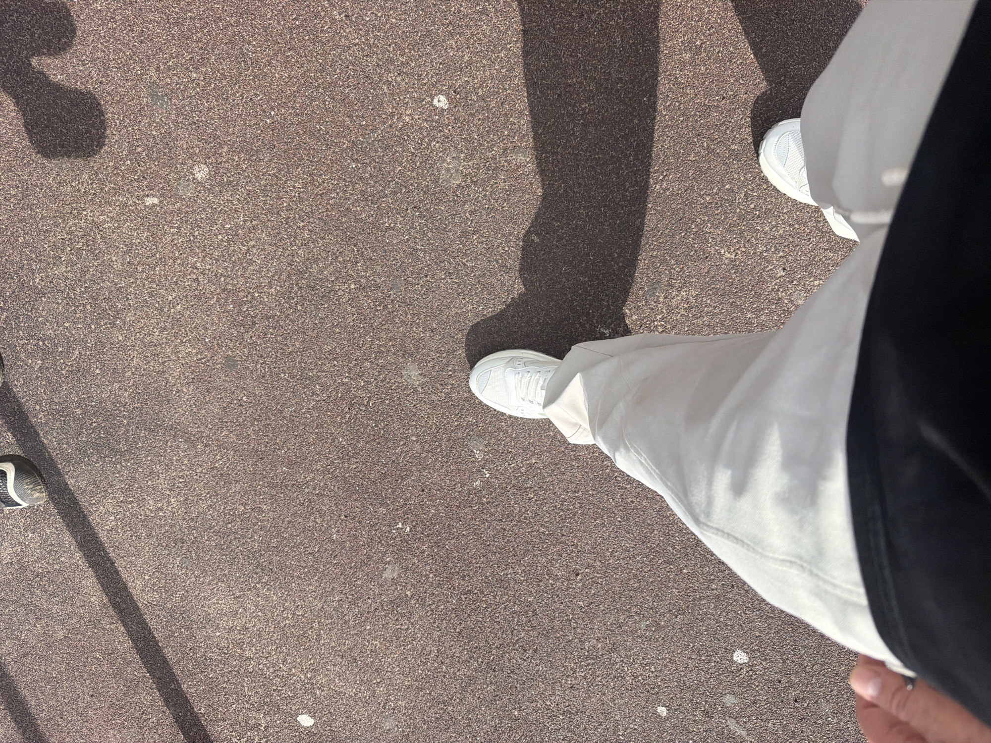 The image shows a person's legs and feet as they walk on a textured, reddish-brown surface, likely asphalt or a similar outdoor material, casting a distinct shadow. The person is wearing light-colored pants and white athletic shoes. A portion of what appears to be a black garment is visible at the bottom of the frame, suggesting the person is wearing a shirt or jacket. The scene is outdoors, with hints of sunlight creating the shadow. There are no discernible landmarks or architectural elements that would specifically indicate Chaikovsky, Russia. The image is a close-up, low-angle shot focusing on the person's lower body and the ground they are walking on.