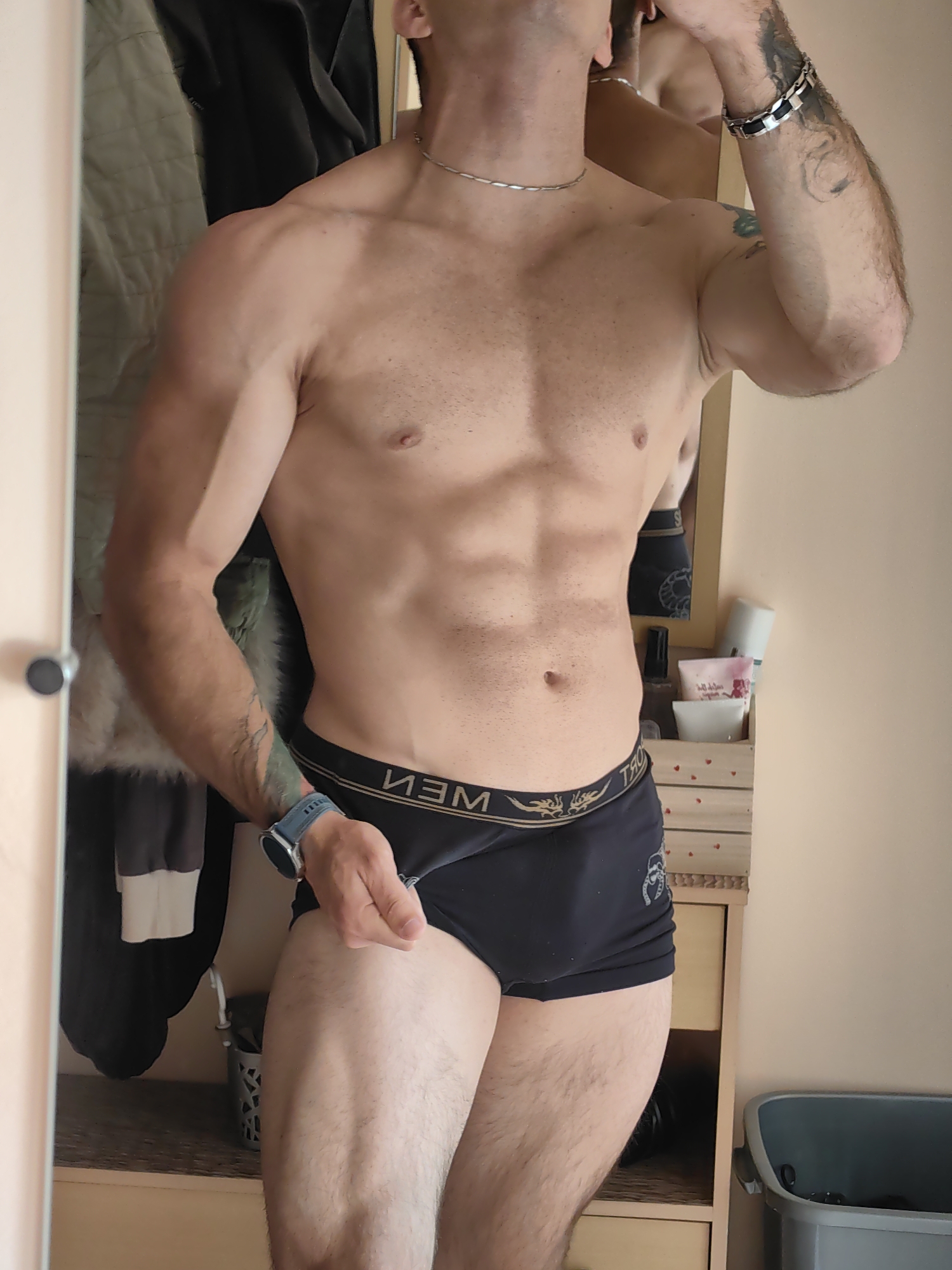 A man is standing in front of a mirror, with his torso and upper thighs visible. He is wearing black boxer briefs with a gold band that has a dragon design on it. He has a watch on his left wrist. His body is toned and muscular. There are tattoos on his left arm. In the background, there is a closet with clothes hanging, a dresser with some toiletries on top, and a trash can. The lighting suggests an indoor setting.