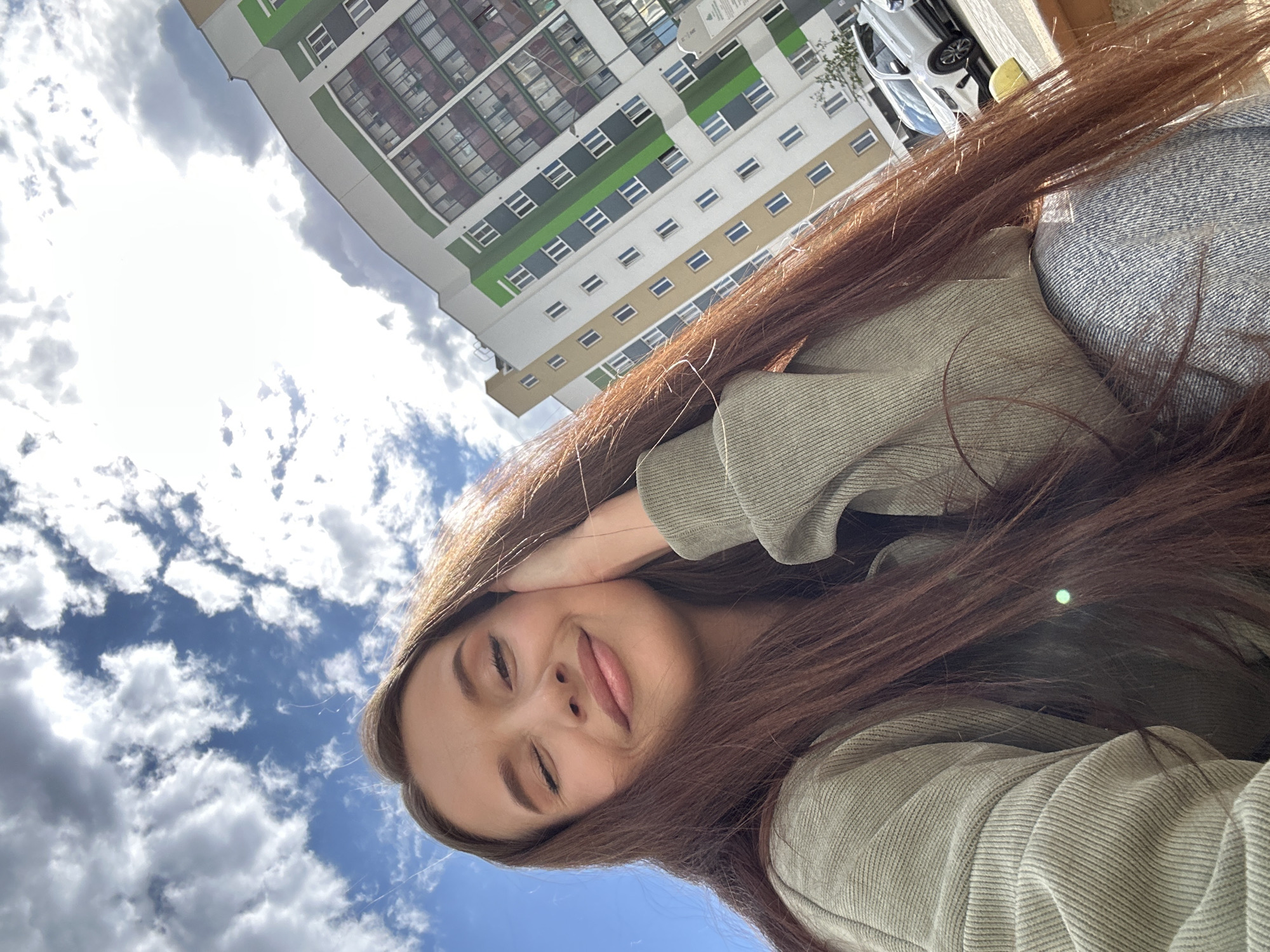 The image appears to be a selfie taken by a young woman in Yekaterinburg, Russia. She has long, brown hair and is wearing a light green sweatshirt and a blue denim jacket. Her eyes are closed, and her lips are slightly parted. Her left hand is raised and touching her head. The background shows a partly cloudy sky and a multi-story apartment building with green, gray, beige, and white facade sections. A white car is partially visible next to the building.