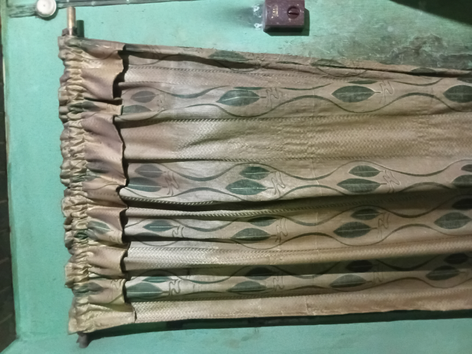 A tan and green patterned curtain hangs in front of a teal-colored wall. The curtain has a pleated top and a repeating leaf design. To the right of the curtain, mounted on the wall, is a small brown electrical box with a round dial. The wall appears textured and shows some signs of wear. The scene suggests an interior setting, likely a room in Nkpor, Nigeria.