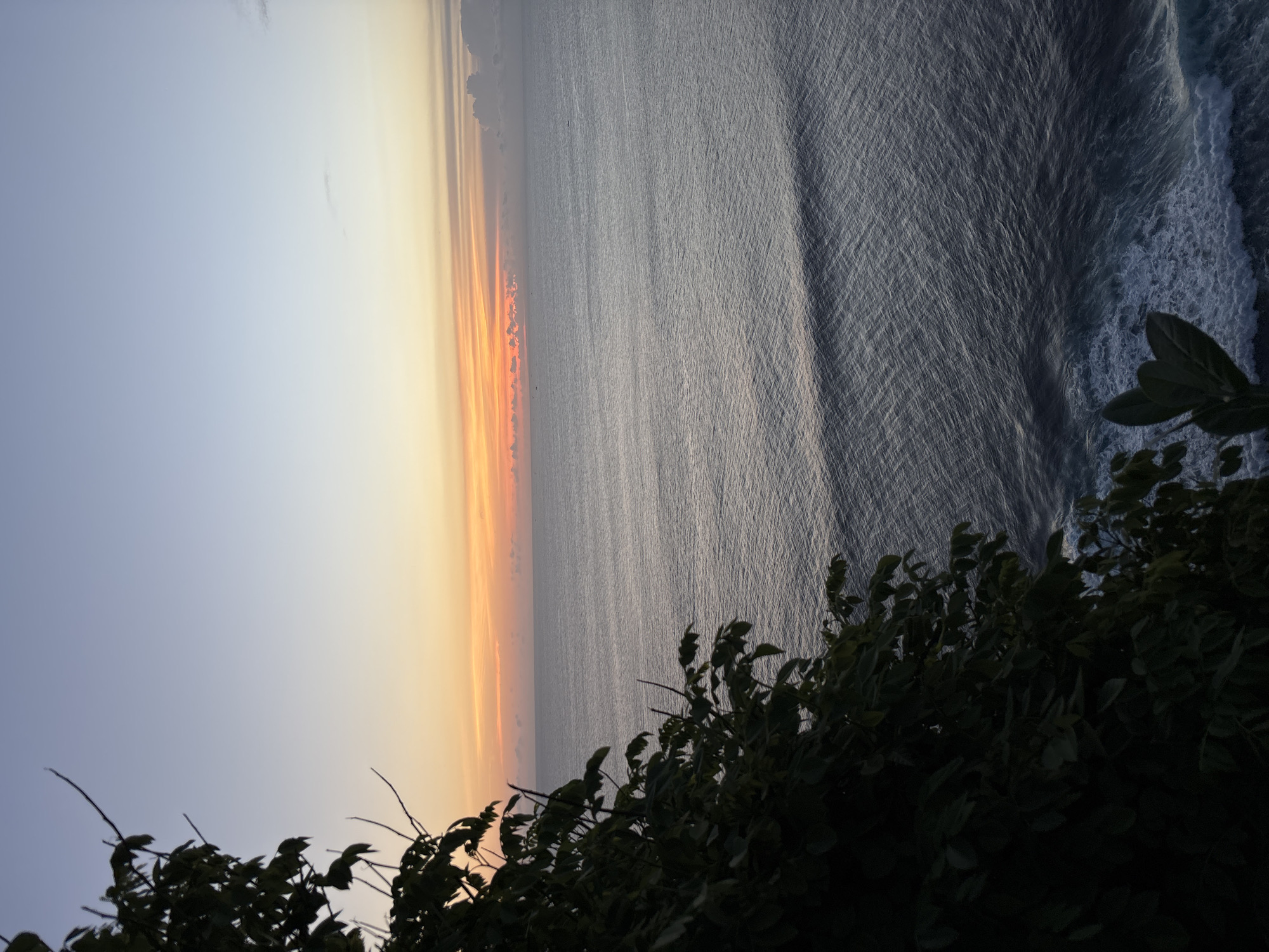 This image shows a view of the ocean at sunset in Pecatu, Indonesia. The sky transitions from a pale blue to a soft orange and yellow gradient at the horizon. A sliver of darker orange hints at the sun's position. The ocean is calm with gentle waves, and its surface reflects the colors of the sky. Some whitewater is visible at the right edge, indicating the approach of waves near the shore. Dark green vegetation, likely foliage growing on a cliff or hillside, occupies the lower half of the frame. The photo is taken from an elevated perspective.