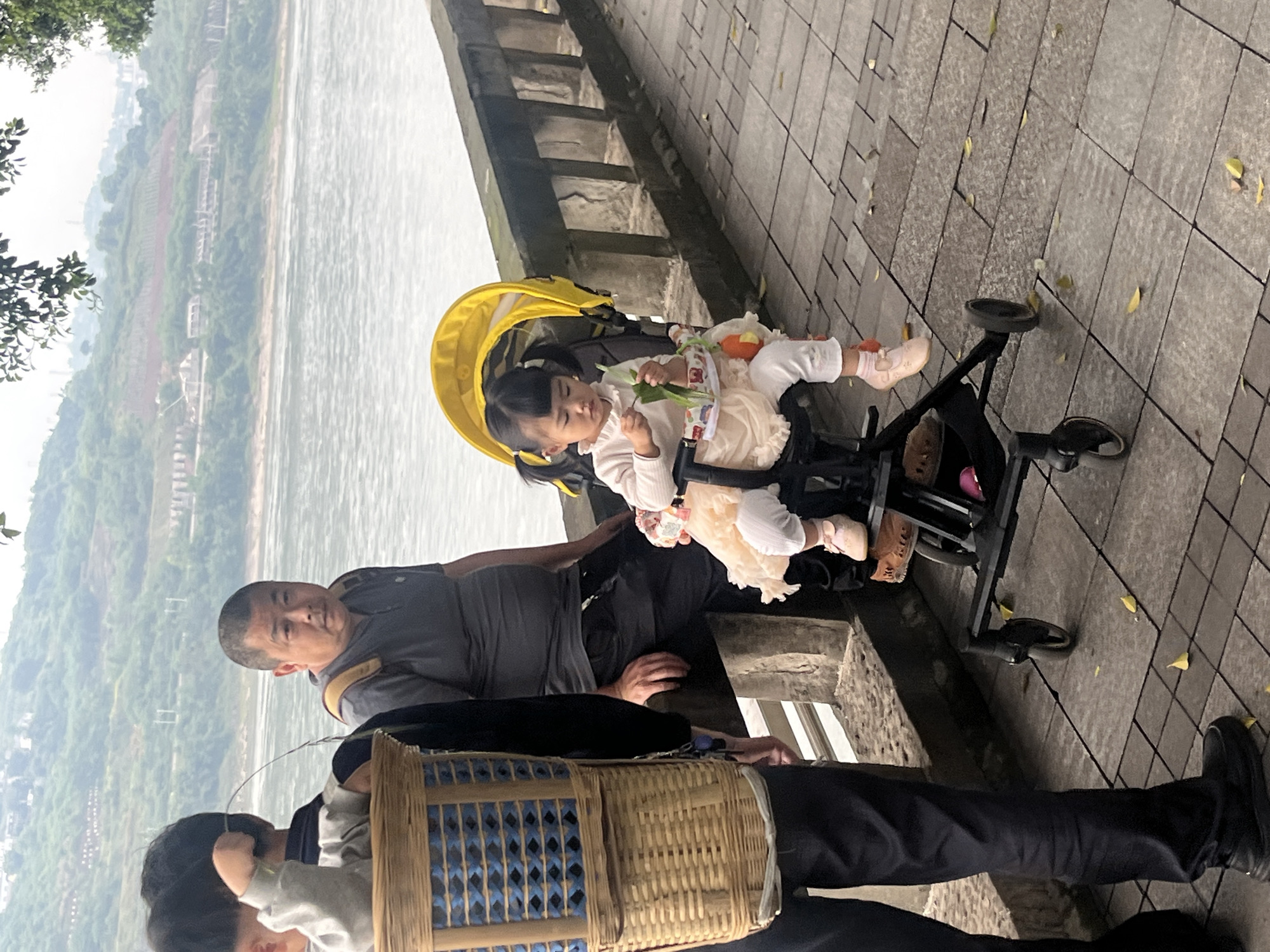 A family outing is captured at a scenic location in Jiangjin, China. In the foreground, a young child sits in a yellow-hooded stroller. The child is fair-skinned with dark hair styled in pigtails. A man with short, graying hair and a gray t-shirt is next to the stroller. Further down, a second man with dark hair wears black pants and holds a woven basket. The scene is set against the backdrop of a wide, river or lake with trees and hills. The location appears to be a pedestrian walkway, indicated by the stone-paved ground.