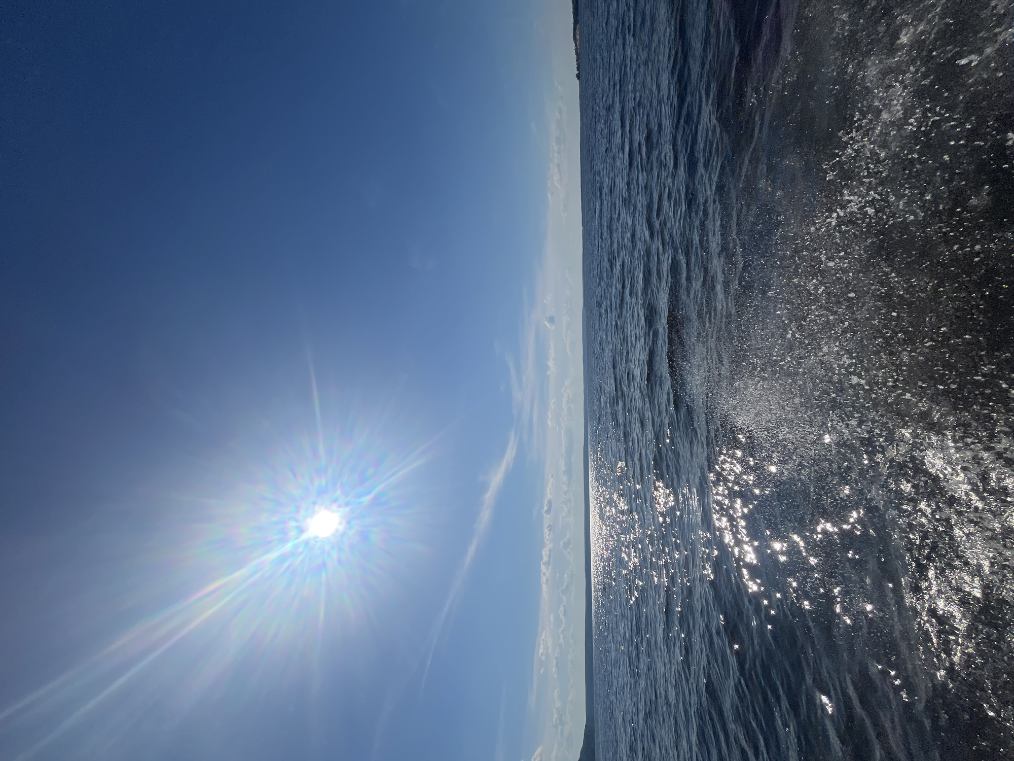 The image depicts a bright, sunny day over a body of water, likely a lake or sea, given the distant landmass visible on the horizon. The sun is intensely bright, casting a prominent lens flare with a rainbow effect across the upper left quadrant of the sky, which is a clear, deep blue. The water is dark blue with whitecaps and sparkling reflections of the sun. In the foreground, on the right side, a splash of water is visible, suggesting movement, perhaps from a boat or wave action. The environment is natural and serene, with a clear sky and calm water. The location is identified as Siukterka, Russia.