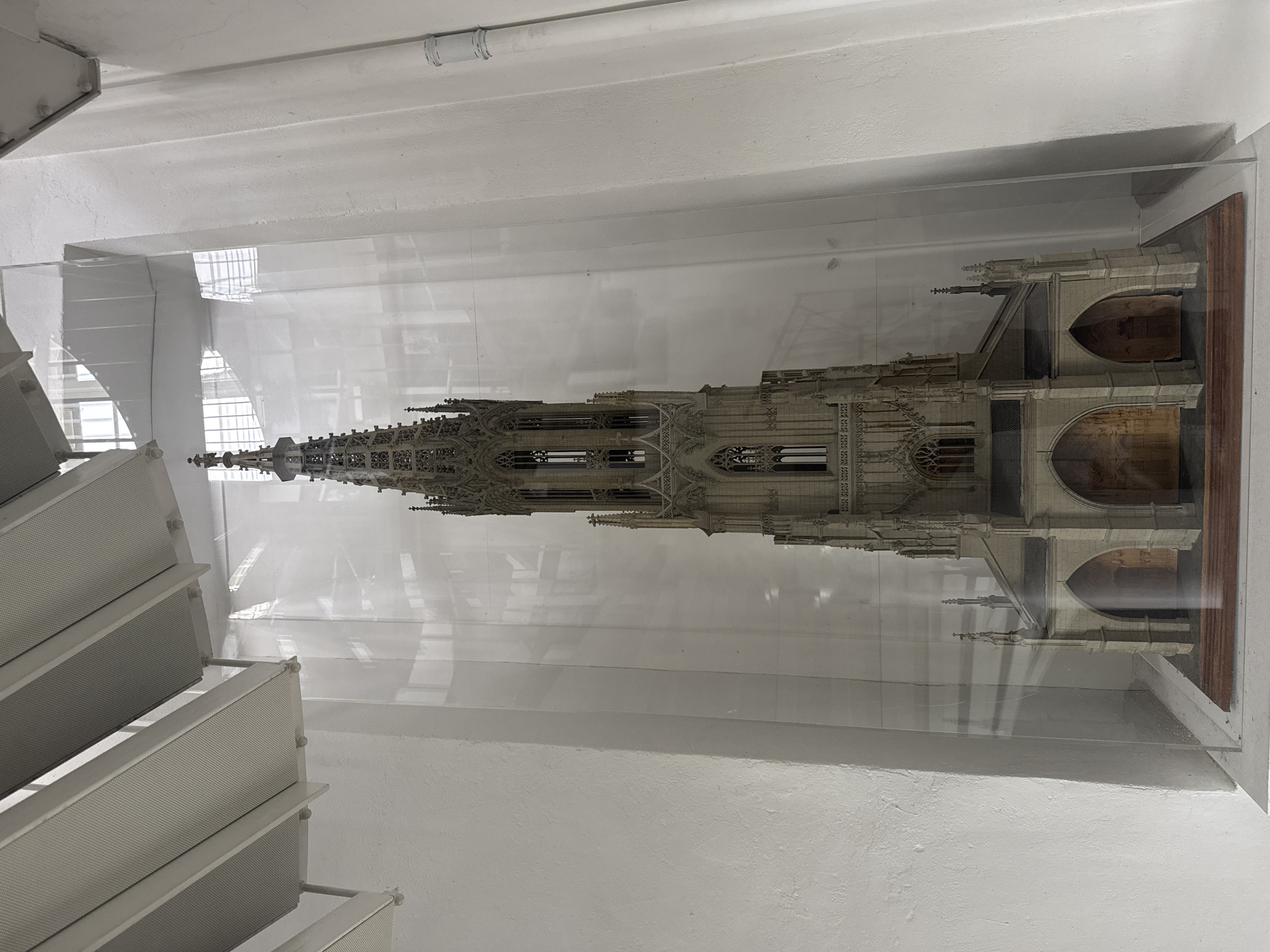 A vertical, eye-level, medium shot captures a detailed architectural model of a cathedral spire displayed inside a clear acrylic case. The model is intricately designed with Gothic architectural elements, including pointed arches, ornate carvings, and numerous spires. It appears to be made of a light-colored material, possibly plaster or resin, and is illuminated from above, casting subtle shadows. The model is positioned horizontally within the case, extending from the left to the right of the frame.
The setting is indoors, with white, textured walls and a white ceiling visible in the background and foreground. A section of a modern, angular white staircase or tiered structure is present in the lower left quadrant of the image, suggesting the model is on display in a museum, exhibition space, or public building. The acrylic case reflects light, creating a hazy effect and obscuring some of the finer details of the model and the background. The overall impression is one of a well-preserved or newly constructed model, highlighting the craftsmanship and grandeur of Gothic architecture. No people are visible, and the scene suggests a static display rather than an active event. The time of day and weather are not discernible from the image. There is no visible text. The location context is Bern, Switzerland.