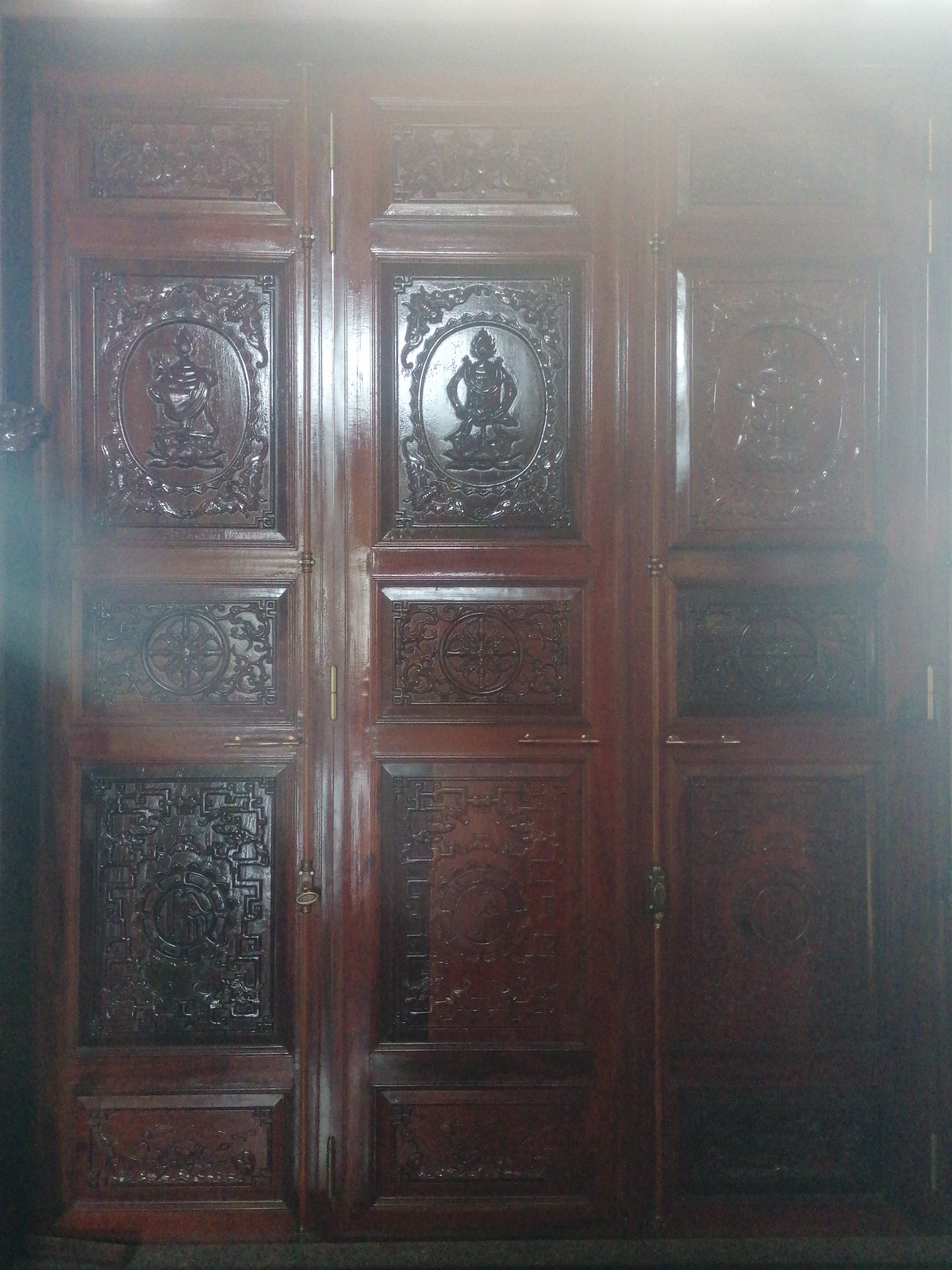 The image shows a close-up of a large, ornate, dark wooden door in Da Lat, Vietnam. The door is divided into six panels, three on each side, with intricate carvings. The top panels on each side feature circular medallions with depictions of figures, possibly deities or historical characters, seated on lotus flowers. The middle panels have square medallions with decorative floral or geometric patterns. The bottom panels are rectangular and also feature detailed carvings of what appear to be scenes or motifs. The door is made of a rich, dark wood with a glossy finish, suggesting it is antique and well-maintained. The lighting in the image is bright and diffused, with a strong highlight on the top portion, which obscures some of the finer details and suggests it might be daytime with sunlight entering the space. There are no people visible in the image. The context suggests this is likely the entrance to a temple, historical building, or a traditional home in Da Lat, a city known for its French colonial architecture and numerous pagodas and historical sites.