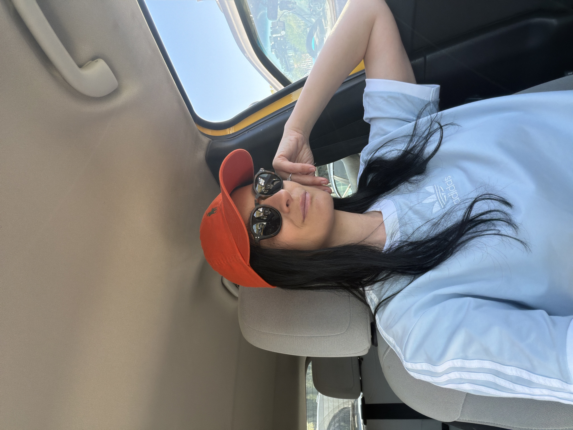 A person is captured in a close-up, horizontal selfie taken inside a car in Tbilisi, Georgia, during bright daytime. The individual, appearing female, has long dark hair and is wearing an orange baseball cap and dark sunglasses. She is dressed in a light blue t-shirt with the white "adidas" logo and text visible on the chest, along with three white stripes on the sleeves. Her left arm is bent, with her hand gently touching her face near the sunglasses. She is reclining against a light gray car headrest, likely in the passenger seat. The car's light-colored headliner and a grab handle are visible above. Through the car window, a clear blue sky is visible, along with reflections in the sunglasses that suggest an outdoor urban or suburban setting with buildings and trees. The car's window frame appears to have a distinct yellow edge.