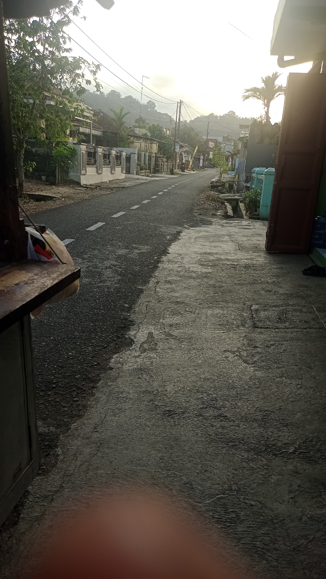 This is a street-level, medium-angle shot taken in Pakemitan, Indonesia, likely during the early morning or late afternoon. The scene depicts a paved road with white dashed lines indicating lanes, curving towards a hazy, tree-covered hillside in the distance. The sky is bright and overcast, suggesting diffused sunlight.
On the left side of the road, there are several houses with tiled roofs and decorated walls. A large tree with green foliage stands prominently in the foreground on the left. A wooden structure, possibly a stall or counter, with some bags on it, is partially visible in the lower left corner.
The right side of the road features a concrete walkway with some cracks and patches. Several containers, some blue and green, are placed near a dark red, open door or gate. A palm tree is visible further down the road. A pair of black sandals rests on the ground near the containers. Electrical wires stretch across the scene, supported by poles. The overall impression is of a quiet, residential street in a rural or semi-rural area.
