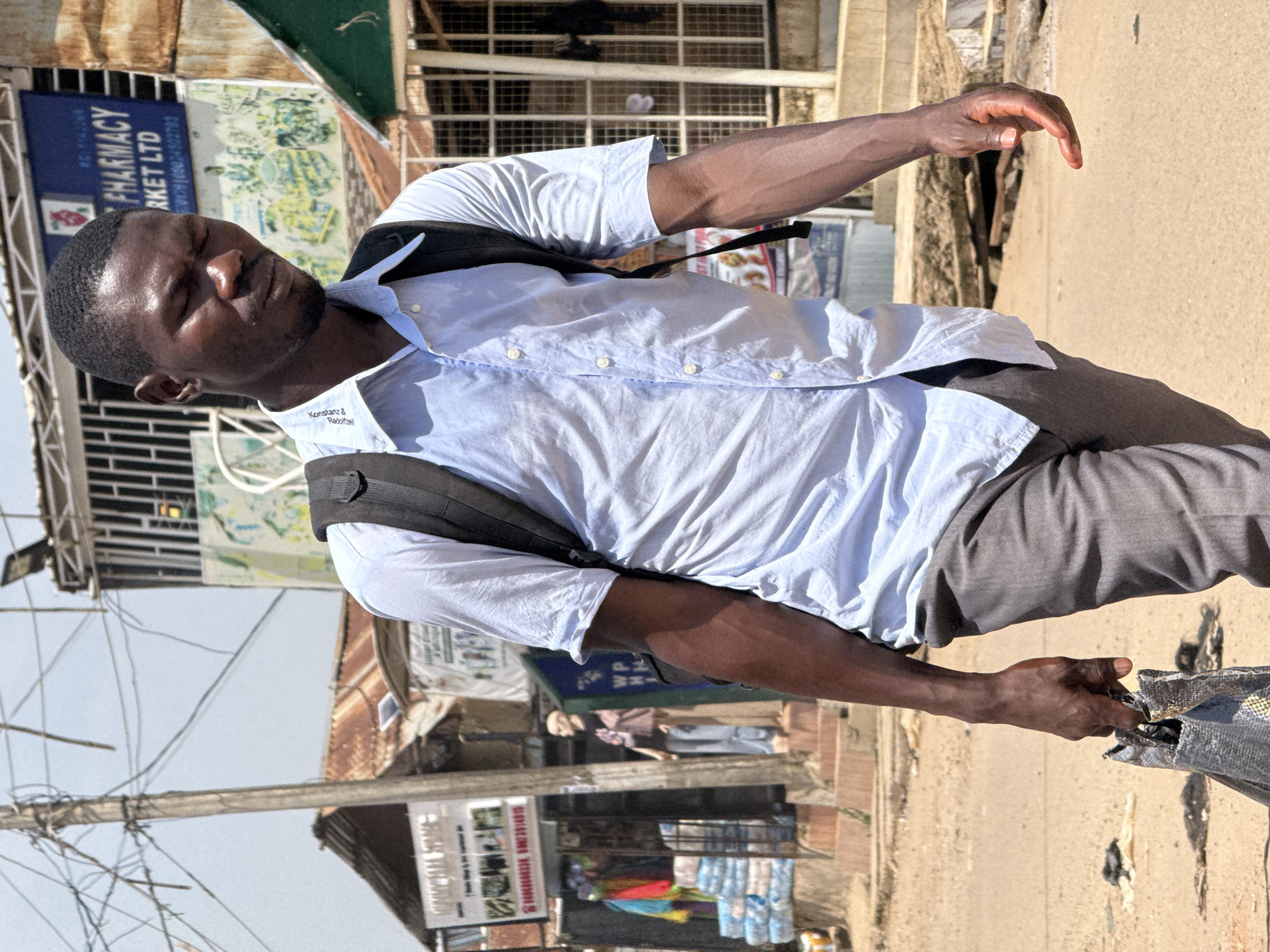 A man, appearing to be in his late 20s or early 30s, walks on a dusty street. He is wearing a light blue button-down shirt, grey trousers, and a black backpack. He carries a crumpled grey bag in his right hand. His eyes are partially closed, and his head is tilted downwards. The background shows a bustling street scene with shops and market stalls. A sign for "PHARMACY MARKET LTD" is visible, and another sign displays the text "Konstanz & Bagozie". The environment appears to be an urban setting in Abuja, Nigeria, with corrugated metal roofs and overhead electrical wires contributing to the characteristic streetscape. The lighting suggests a bright, sunny day.