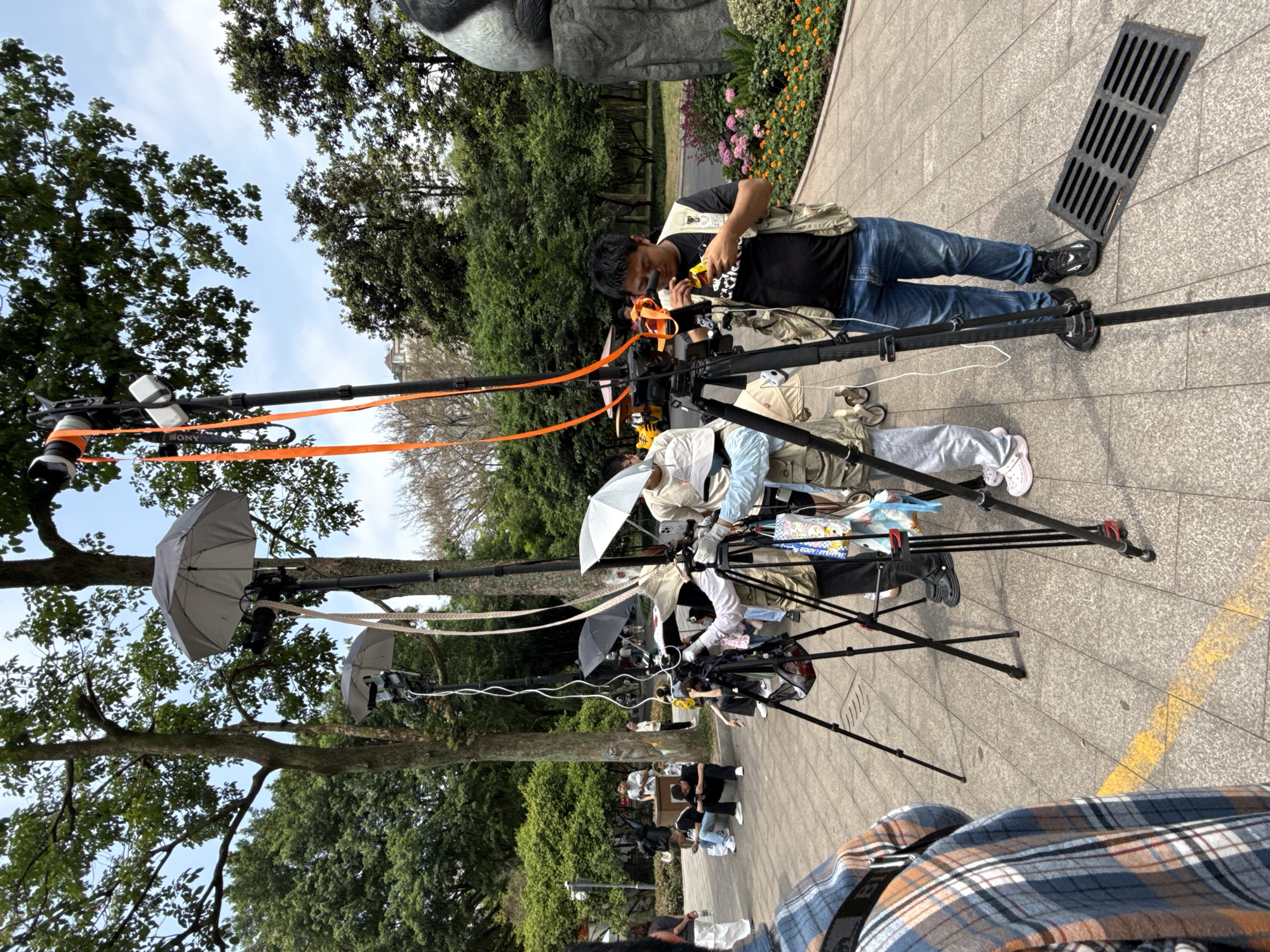 The image shows a film crew preparing for a shoot in Jiulongpo District, China. A man is adjusting equipment on a tall camera rig, while other crew members are working on similar rigs with attached umbrellas. Several people are sitting or standing in the background. The location appears to be a park, with trees and flowers visible. The ground is paved with stone tiles, and there is a drainage grate near the main subject.