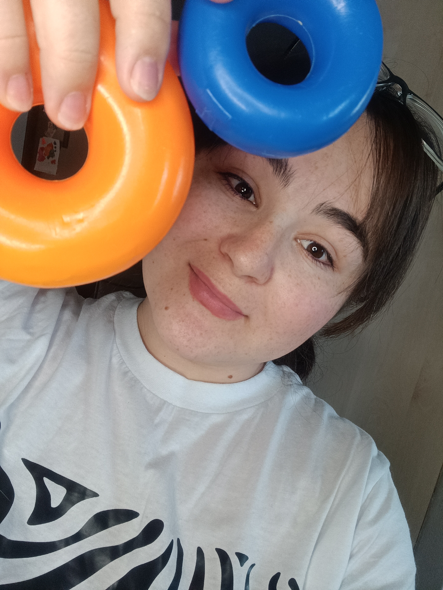 A young woman with brown hair and freckles is holding an orange plastic ring in her left hand and a blue plastic ring above her forehead. She is wearing a white t-shirt with a black zebra stripe design. Glasses are perched on top of her head. The background is a neutral color. The location, Noviy, Russia, is not discernible from the image.