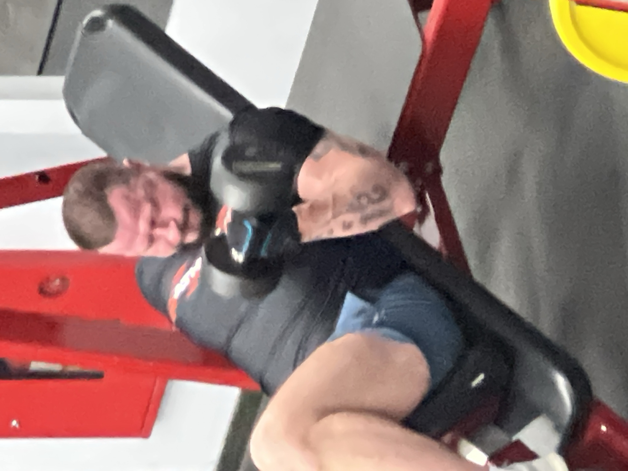 A man is working out on a piece of exercise equipment at a gym. He is positioned face down with his torso and legs supported by pads. He is wearing a black shirt and shorts. The exercise machine is red and black. The gym appears to be indoors with light-colored walls. The location is likely in Khimki, Russia.