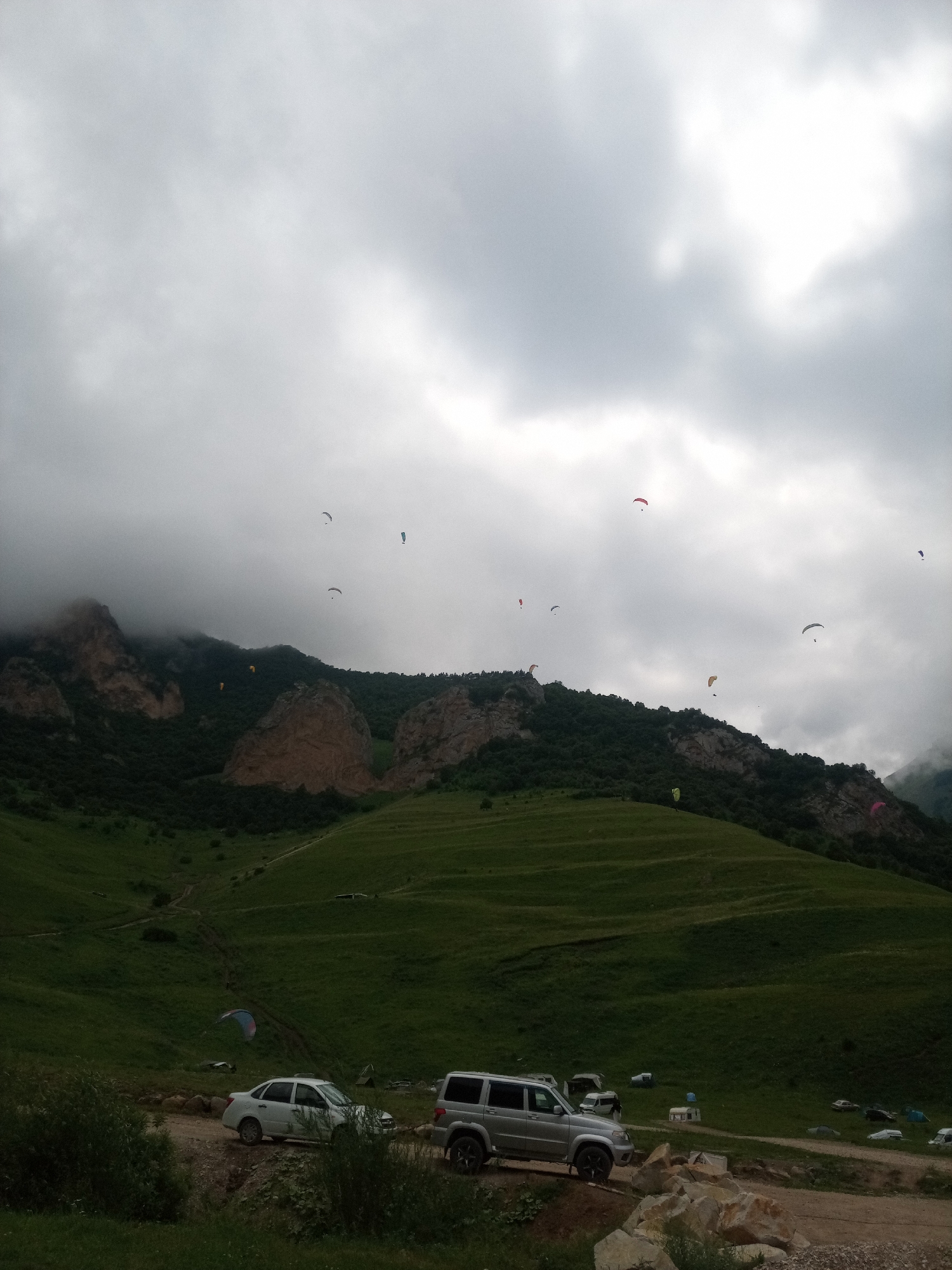 The image depicts a mountainous landscape with several paragliders in the sky. The scene is located in El'tiubiu, Russia. In the foreground, there is a dirt road with two vehicles parked on it, a silver sedan and a silver SUV. The surrounding area is covered in green grass and vegetation. In the background, there are steep, rocky mountains covered in trees. The sky is overcast with clouds. Several paragliders, some with colorful canopies, are visible in the air, appearing to be engaged in recreational activities. The overall impression is one of an outdoor adventure or sporting event taking place in a scenic, natural environment.