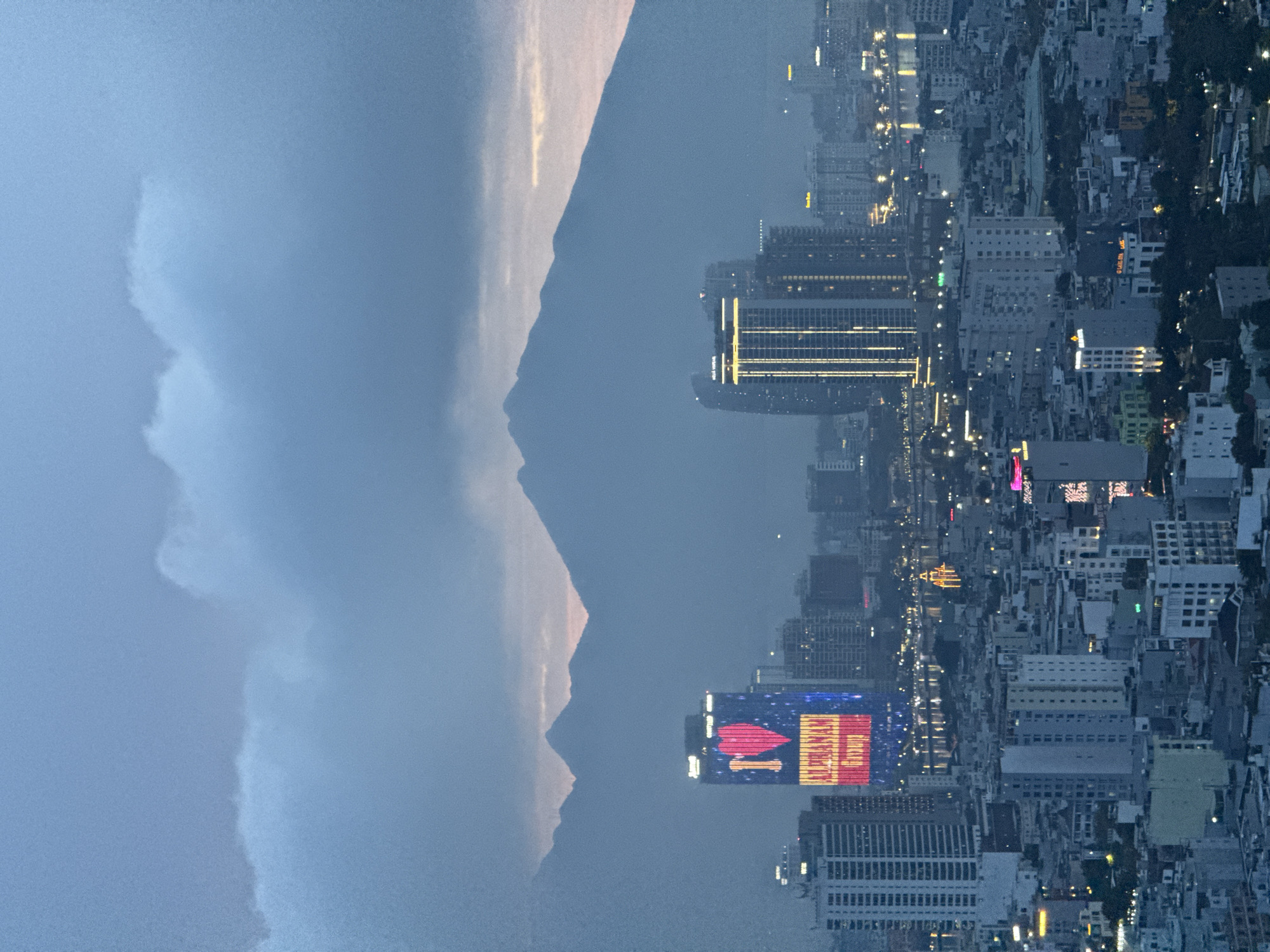 This image captures a panoramic view of Đà Nẵng, Vietnam, at dusk. The city skyline is dominated by numerous high-rise buildings, many of which are illuminated with lights, suggesting the early evening hours. Prominently featured is a tall skyscraper displaying a large, illuminated red heart with the text "I" and "ALPHANAM GROUP" in vibrant yellow and red lights. This building is situated in the foreground, to the left of the frame, and appears to be a significant landmark.
Further into the city, other buildings of varying heights are visible, with windows lit and some displaying subtle architectural lighting. A body of water, likely a river or bay, runs through the cityscape, reflecting the lights of the city and creating a shimmering effect. On the far side of the water, more buildings and lights are discernible, extending the urban landscape.
In the background, a range of majestic mountains forms a dramatic backdrop to the city. Their silhouettes are softened by the hazy atmosphere of dusk and possibly light fog or clouds. The sky is a gradient of muted blues and grays, with hints of soft pink and orange near the horizon, indicating the transition from day to night. Clouds drift across the upper portion of the sky, adding depth and texture to the scene. No people are directly visible in this aerial perspective. The overall impression is one of a bustling, modern city nestled against a natural, mountainous landscape during twilight.