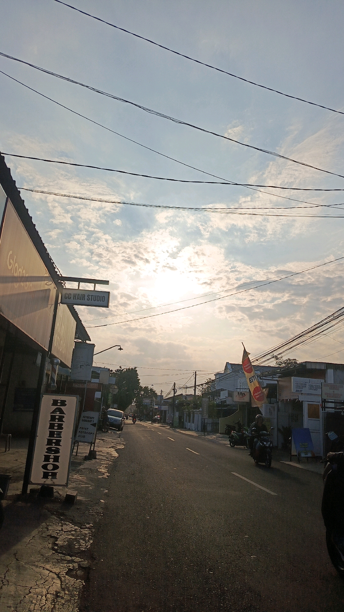 The image depicts a street scene in Ronowijayan, Indonesia, during what appears to be sunrise or sunset, given the low angle of the sun and the warm lighting. The sky is partly cloudy, with the sun partially obscured by clouds. Overhead, a tangle of electrical wires crisscrosses the sky. On the left side of the street, a building with a sign that reads "Glodak" is visible, along with a smaller sign for "CC HAIR STUDIO". Further down the sidewalk, a tall, vertical sign for "BARBERSHOP" stands prominently, with a barber pole at its base. Several other signs and storefronts line the street. A paved road stretches into the distance, with a white line marking the center. A few motorcycles are present, with one rider visible on the road. The overall impression is a typical urban street in a developing area, with evidence of commercial activity and daily life.