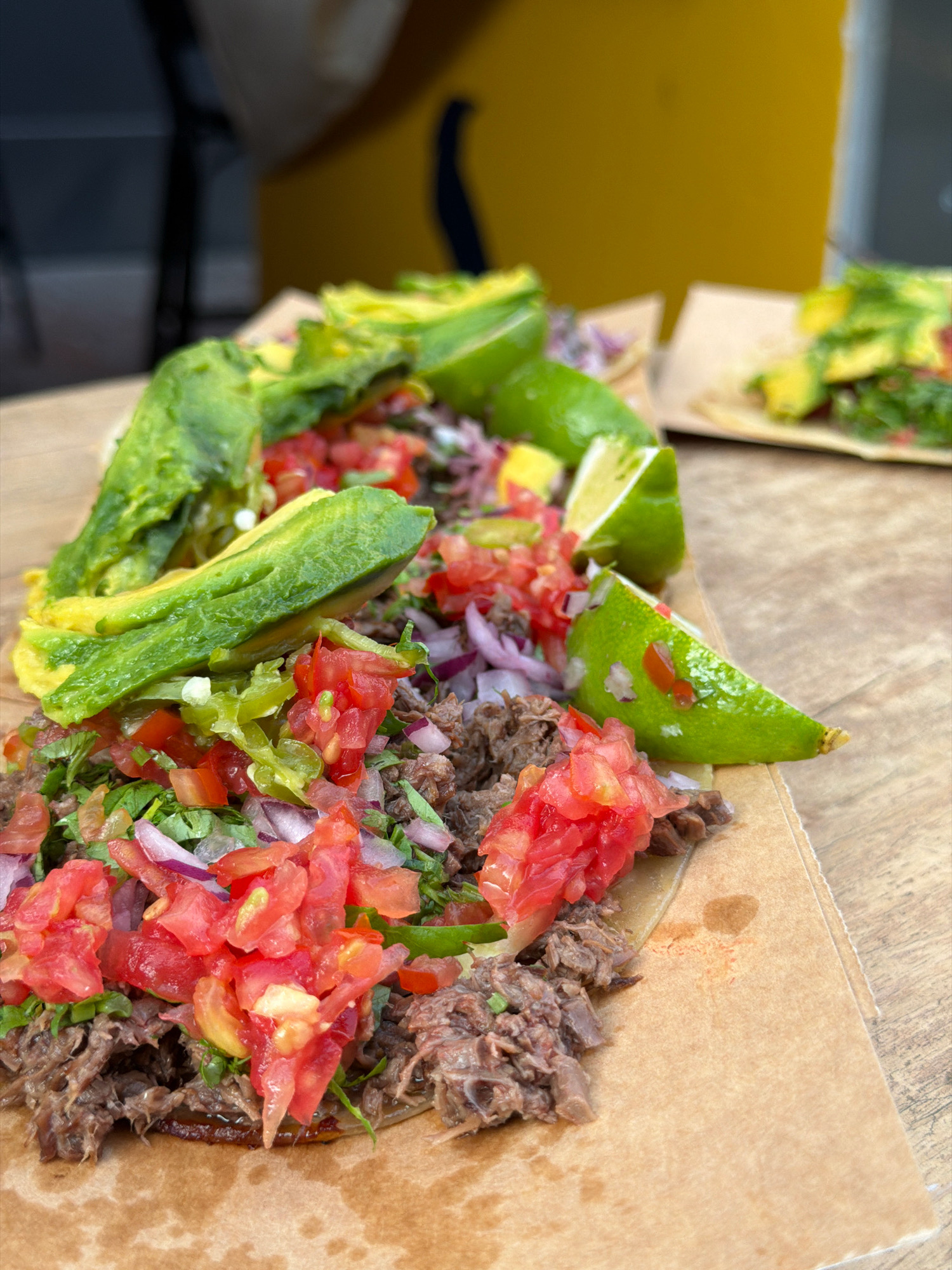 This is a close-up, eye-level shot of two shredded meat tacos with avocado and pico de gallo, served on a piece of brown paper placed on a wooden table. The tacos are topped with sliced avocado, diced tomatoes, finely chopped red onions, and a scattering of green herbs. Lime wedges are visible on the side of the tacos. The background is blurred, showing a yellow and dark blue object, possibly part of a vendor's stand, suggesting an outdoor food setting. Based on the overall composition and presentation, this scene is likely from a food stall or market in Tel Aviv, Israel.