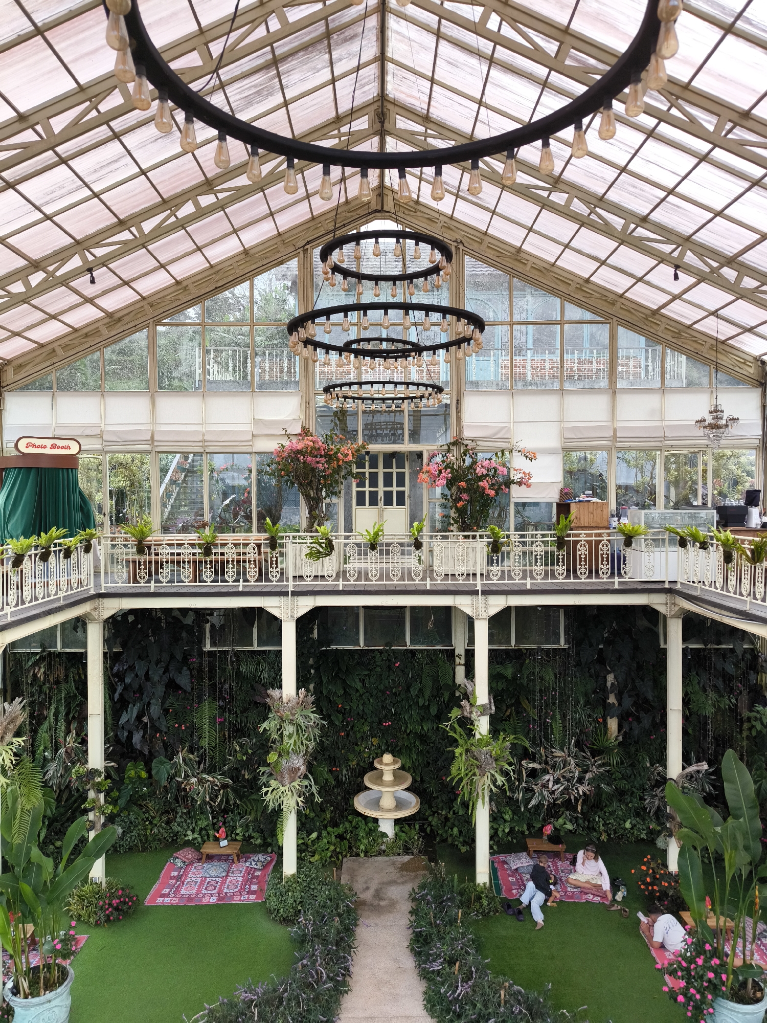 Captured from an elevated vantage point inside a spacious greenhouse-like building, the scene reveals a multi-tiered, lush interior. The structure boasts a glass-paneled roof and walls, allowing ample natural light to permeate the space. Massive circular chandeliers with numerous bulbs hang from the ceiling. A second-story balcony lined with plants overlooks the ground floor, which features artificial turf and a variety of plants and ferns. On the ground level, there are red rugs laid out with small tables, suggesting a lounge area. People are sitting and reclining on the rugs, engaging in leisurely activities. A stone pathway leads to a small fountain in the center of the ground floor. The "Photo Booth" sign is visible on the upper balcony. The location is in Candikuning, Indonesia.