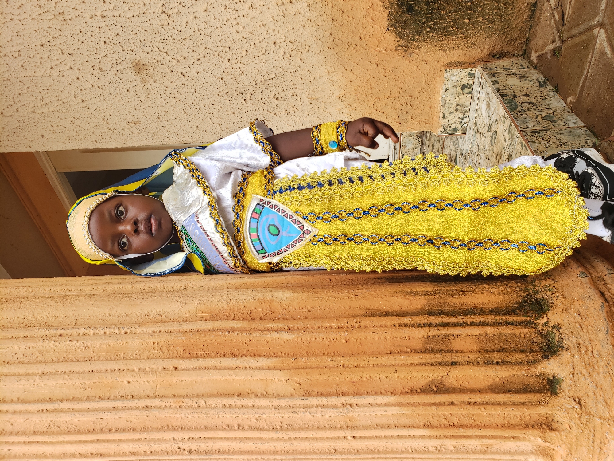 A young child, possibly a girl, is standing outdoors next to a large, textured, orange pillar. The child is wearing a traditional outfit consisting of a white robe with elaborate gold and blue trim, a yellow patterned skirt, and a yellow headscarf adorned with beaded trim. The outfit has intricate embellishments, including a decorative panel on the chest featuring a blue eye-like symbol within a triangular design. The child's left arm is visible, wearing a yellow bracelet with a blue stone. The child is looking towards the viewer with wide eyes.
The background consists of a rough, textured, tan-colored wall and a series of tiled steps. The pillar has vertical grooves and appears to be made of a material like terracotta or concrete. The environment suggests an outdoor setting in a residential or public area, possibly in Sabon Tasha, Nigeria, given the architectural style and context. The lighting indicates daytime. The overall scene captures the child in ceremonial or traditional attire.
