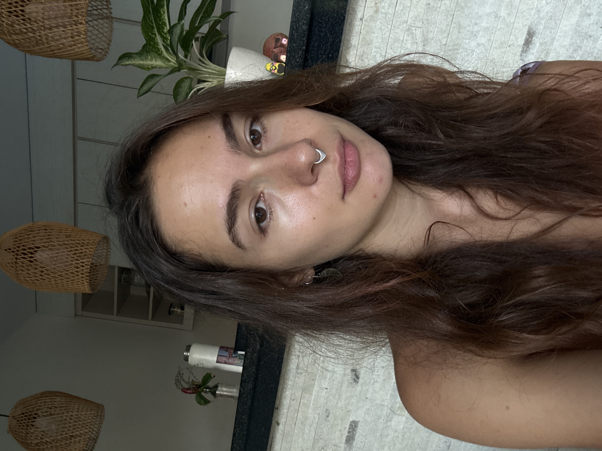 A young woman with medium-length, wavy brown hair is facing the camera. She has a light complexion, brown eyes, and a piercing in her left nostril. She is wearing small hoop earrings. The background includes a countertop, a plant in a white pot, and woven lampshades hanging from the ceiling. The setting appears to be a residential interior, possibly a kitchen or dining area. There is no discernible evidence indicating a location in Buduk, Indonesia.