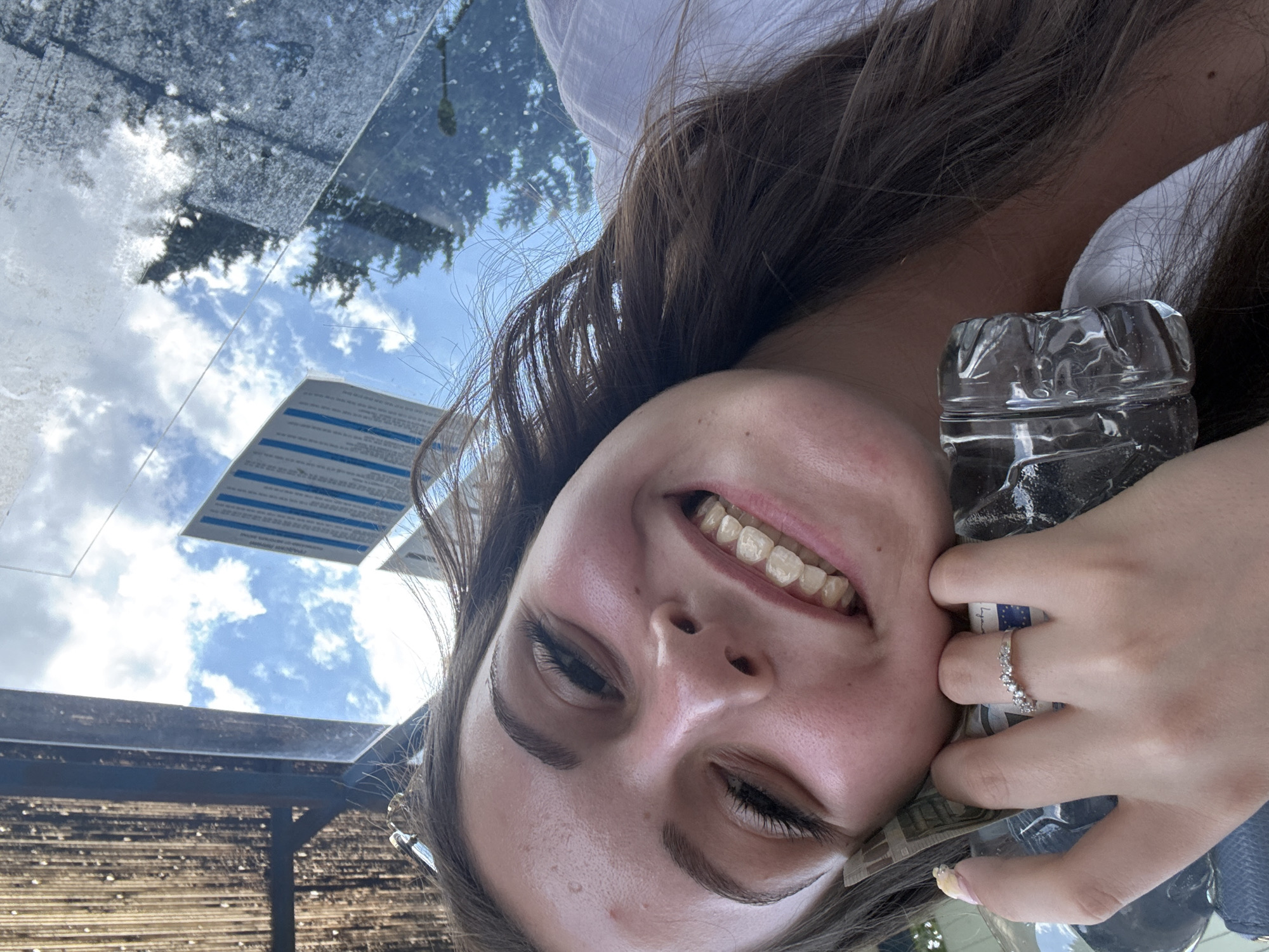 A young woman with brown hair and fair skin is taking a selfie outdoors. She is smiling broadly, revealing her teeth. Her right hand is visible, resting on what appears to be a plastic bottle and some banknotes. She is wearing a ring on her left hand, which is also visible. The background shows a sky with clouds and some reflections, possibly from a window or a glass structure. A tall, blue and white sign is visible in the distance. The overall impression is a casual, everyday moment captured in what appears to be an urban setting. There is no specific indication of the city of Burgas, Bulgaria within the image itself.