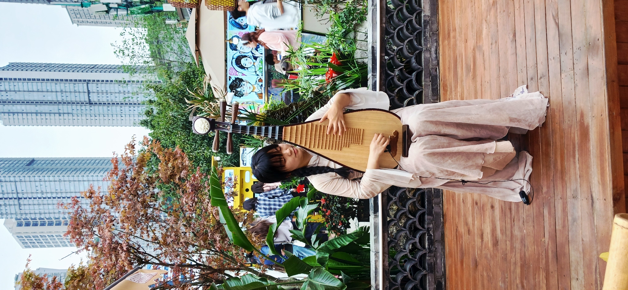 The image depicts a woman playing a pipa, a traditional Chinese lute, in an outdoor setting, likely in Yuzhong District, China. The woman has long black hair styled in a braid and is wearing a pale pink dress. She is seated on a wooden platform. Her left hand holds the neck of the pipa while her right hand plucks the strings.
Behind the woman, there is an array of plants and trees, including banana trees and trees with red-brown leaves. People are standing in the background, some of whom are looking at a painted display of Asian characters. Tall buildings are visible in the far background, indicating an urban environment. A black ornate fence separates the woman from the backdrop. A black microphone is clipped to the dress in front.